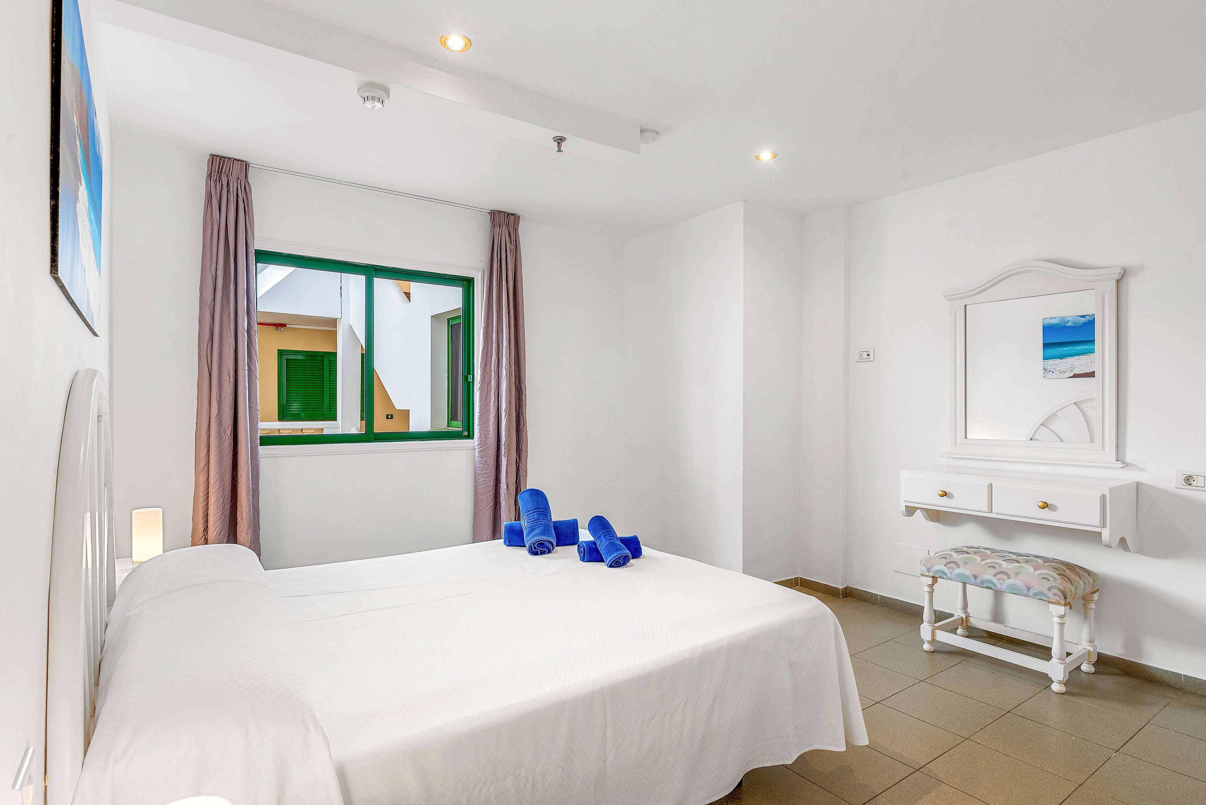 A clean, minimalist bedroom features a double bed with a white bedspread and neatly folded blue towels on top. The room is bright with white walls, tiled flooring, and a green-framed window showing an exterior building. Soft curtains frame the window, and a small white dressing table with a mirror and patterned stool sits against the wall. Recessed ceiling lights and a bedside lamp provide additional warm lighting, creating a calm and inviting atmosphere.