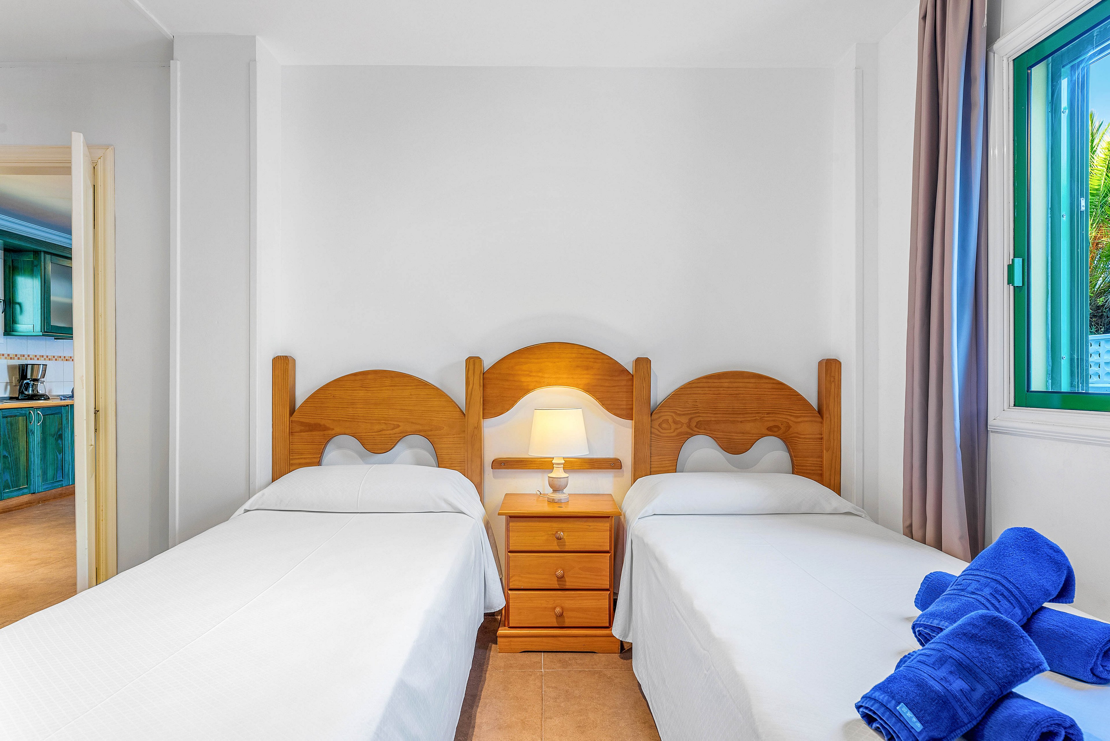A clean, bright bedroom features two single beds with wooden headboards and white bedding. A small wooden nightstand with a lit lamp sits between the beds, creating a cozy focal point. Rolled blue towels are neatly placed on one bed, adding a pop of color to the neutral room. A doorway reveals a green-toned kitchen area, and a window with green trim lets in natural light and a view of greenery outside.