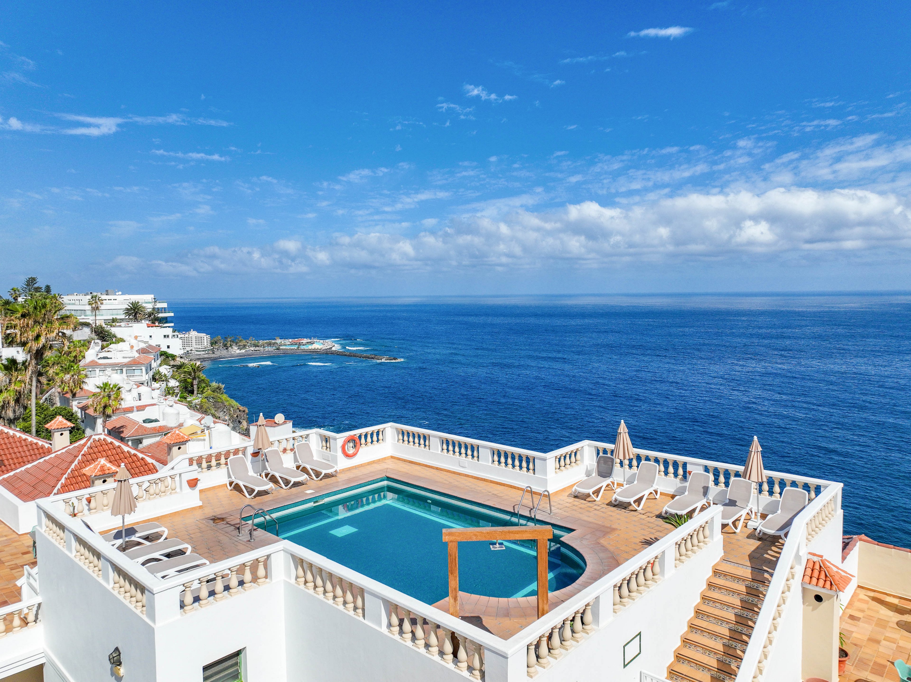 A rooftop swimming pool sits atop a white coastal building with sun loungers arranged around the water. The scene overlooks a vast blue ocean stretching to the horizon under a partly cloudy sky. Nearby terracotta-roofed houses and palm trees line the shoreline, suggesting a warm seaside resort. The bright sunlight and clear weather create a relaxing vacation atmosphere.