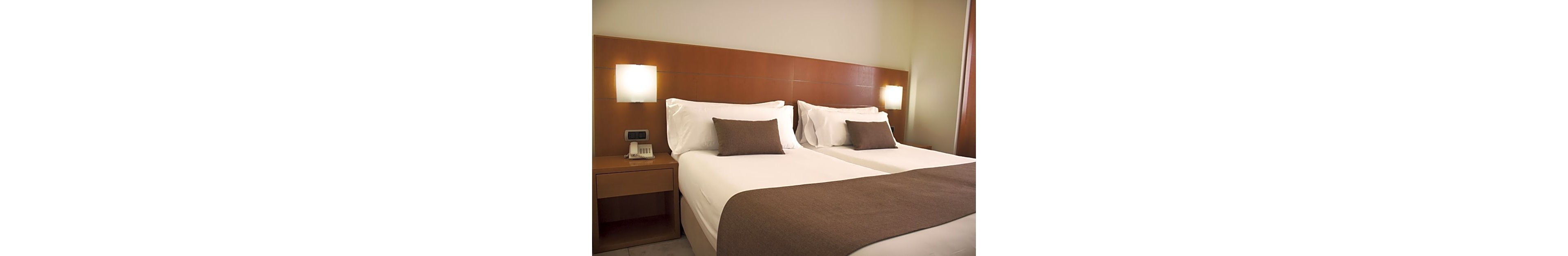 A contemporary hotel room features two neatly made twin beds with white linens and brown throws. A wooden headboard spans the wall behind the beds, creating a warm and minimalist look. Soft wall-mounted lamps provide ambient lighting on either side, with a bedside table holding a telephone and notepad. The overall atmosphere is clean, orderly, and inviting, suitable for business or leisure travel.