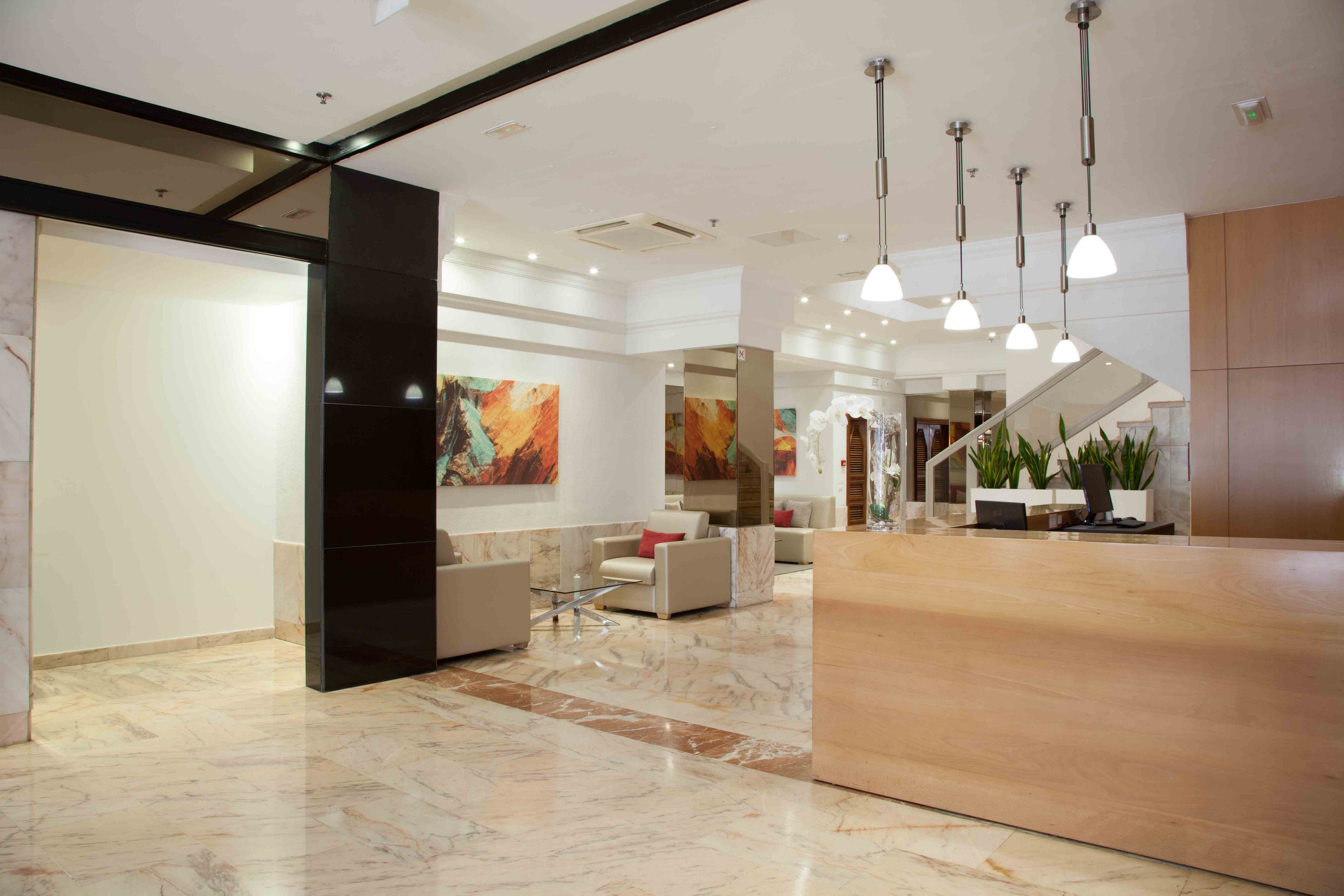 A contemporary hotel lobby features a light wood reception desk and polished marble floors. Neutral-toned sofas and armchairs create a comfortable seating area in the background. Abstract colorful artwork decorates the white walls, adding visual interest. Pendant lights hang from the ceiling, illuminating the clean and minimalist interior space.