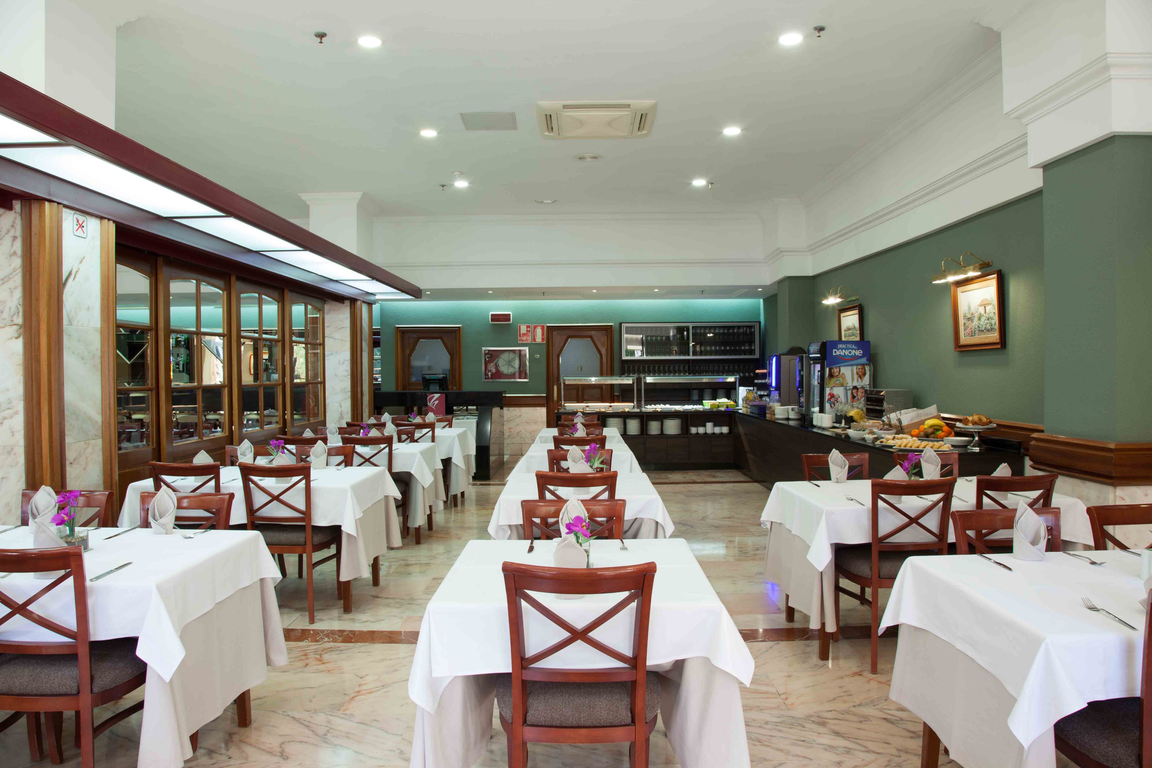 A neatly arranged restaurant dining room features rows of square tables covered with white tablecloths and wooden chairs. The space appears to be part of a hotel or buffet-style venue, with a self-service counter and beverage machines visible at the back. Soft green walls, framed artwork, and glass-front cabinets add a classic, understated decor. The room is brightly lit and empty, creating a calm and orderly atmosphere.