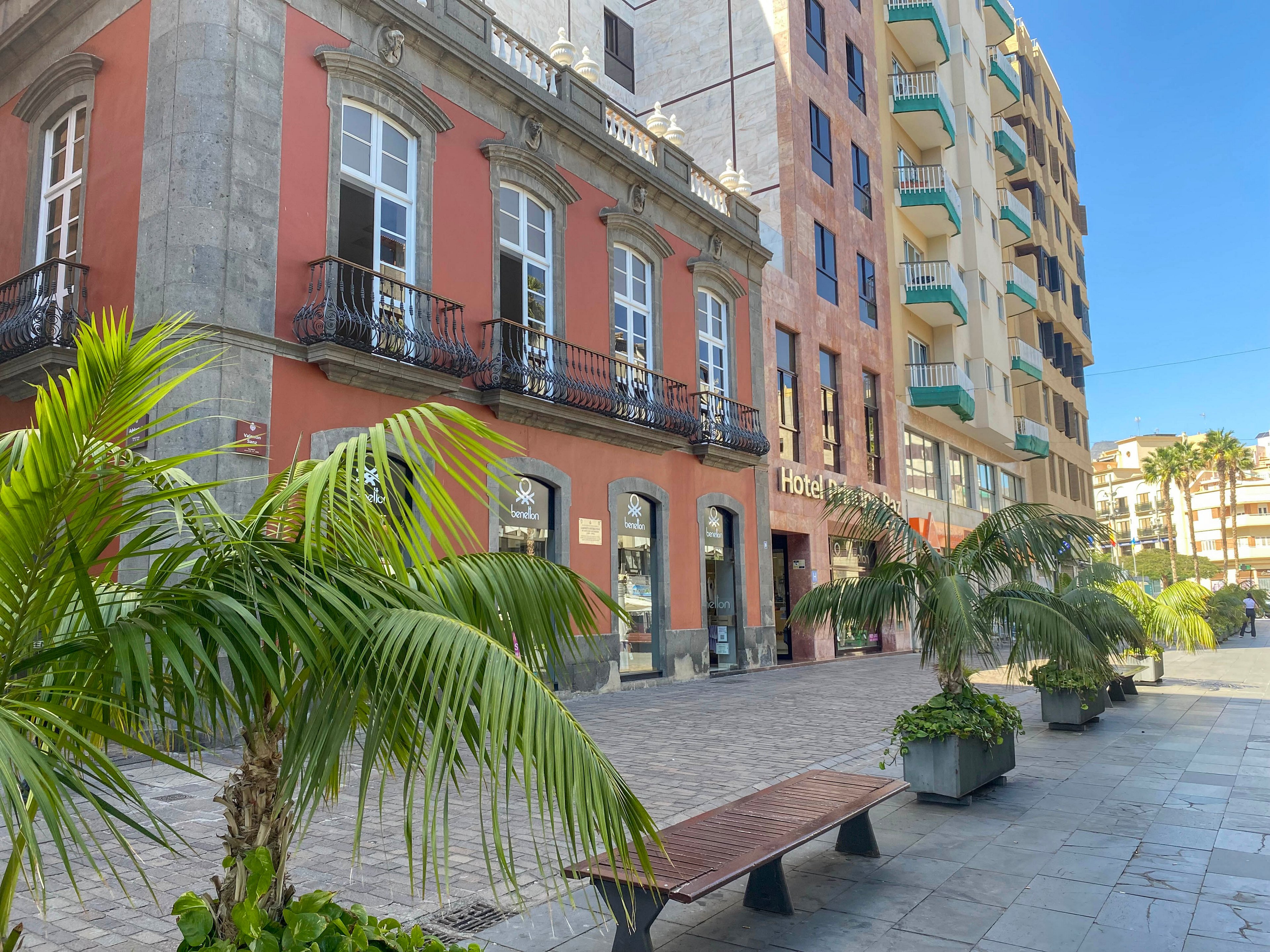 A sunny urban street features a historic red and gray stone building next to a modern hotel. Tall windows with wrought-iron balconies overlook a pedestrian walkway lined with palm trees. Wooden benches and planters create a relaxed public space in front of the hotel entrance. Bright daylight and clear blue sky give the scene a warm, inviting atmosphere.