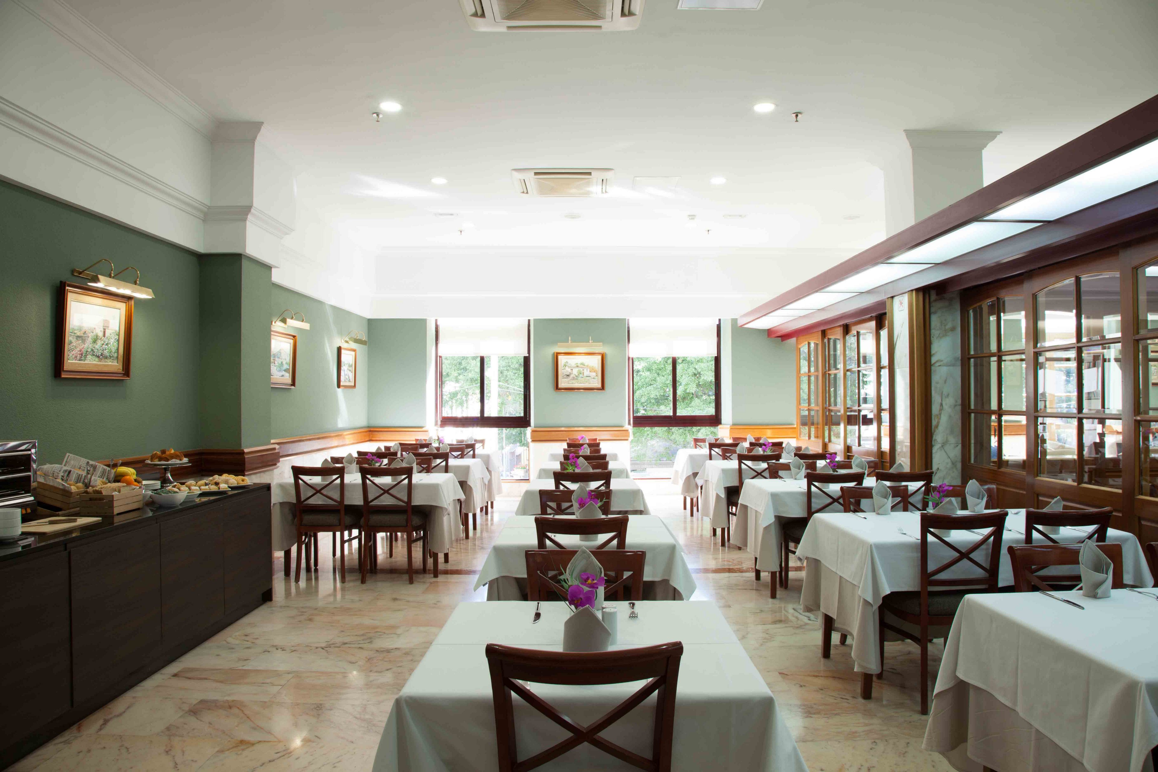 Spacious restaurant dining room with neatly arranged tables covered in white tablecloths. Wooden chairs with cross-back design surround each table, some set with small flower centerpieces. Large windows at the back let in natural daylight, illuminating green walls decorated with framed artwork. A buffet counter lines one side of the room, creating a welcoming setting for breakfast or lunch service.