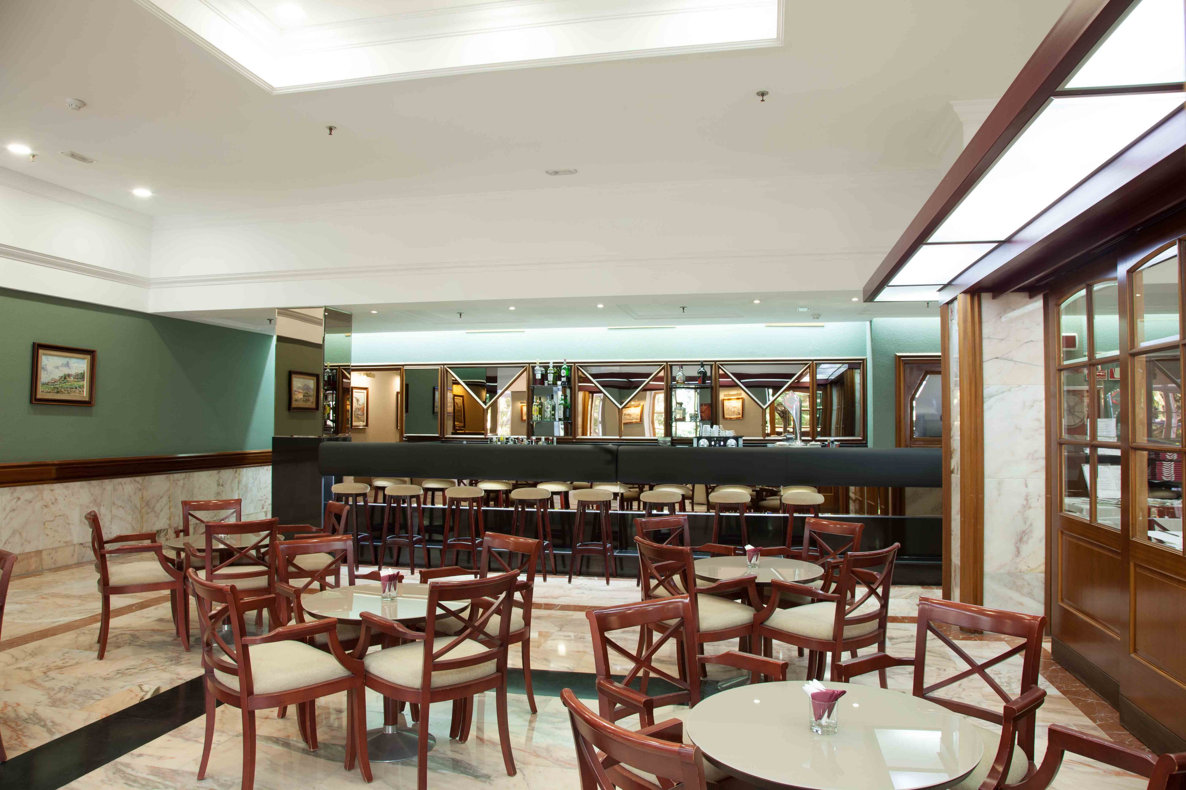 Spacious hotel bar lounge featuring rows of wooden chairs and round tables arranged neatly in front of a long counter. The setting appears clean and modern, with polished floors and a mix of marble and wood finishes. Soft ceiling lighting and large mirrors behind the bar create a bright, open atmosphere. No people are present, emphasizing the calm, orderly interior space.