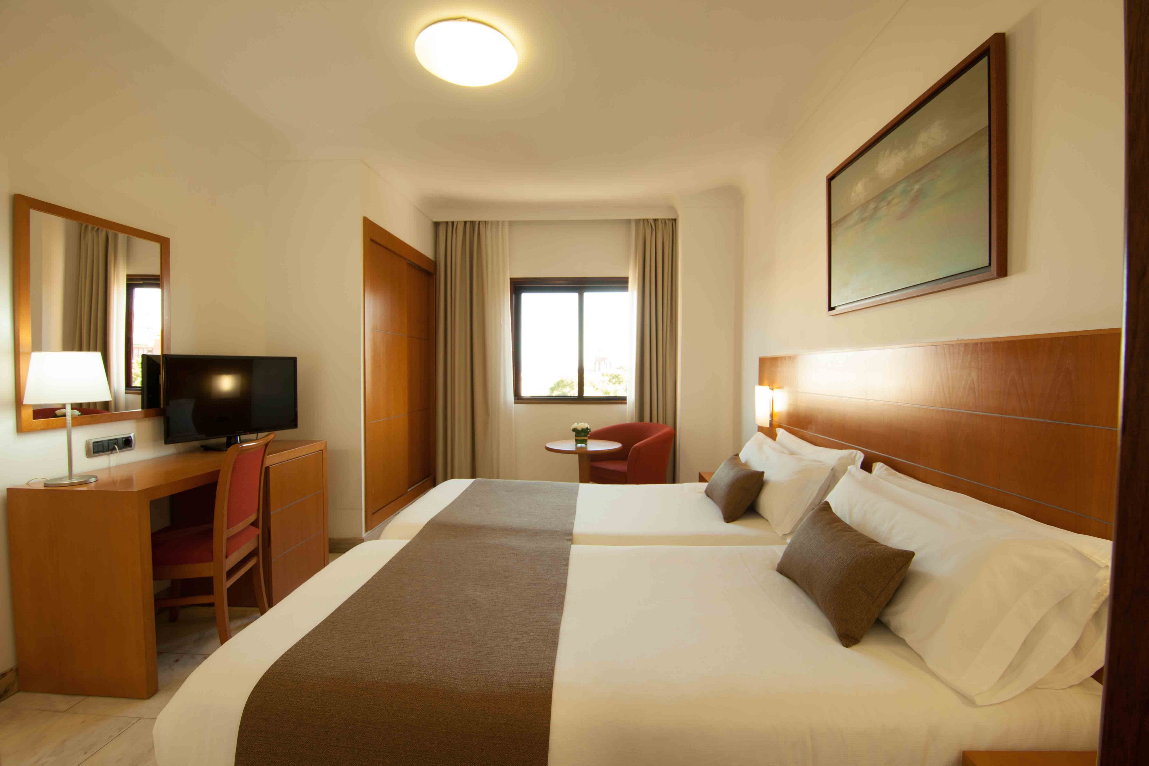 A warm, modern hotel room features two neatly made twin beds with white linens and brown throws. A wooden headboard and matching furniture, including a desk with a flat-screen TV and lamp, create a cohesive design. Natural light enters through a central window, illuminating a small round table and red armchair in the corner. Soft lighting and neutral tones give the space a calm, inviting atmosphere suitable for rest or work.
