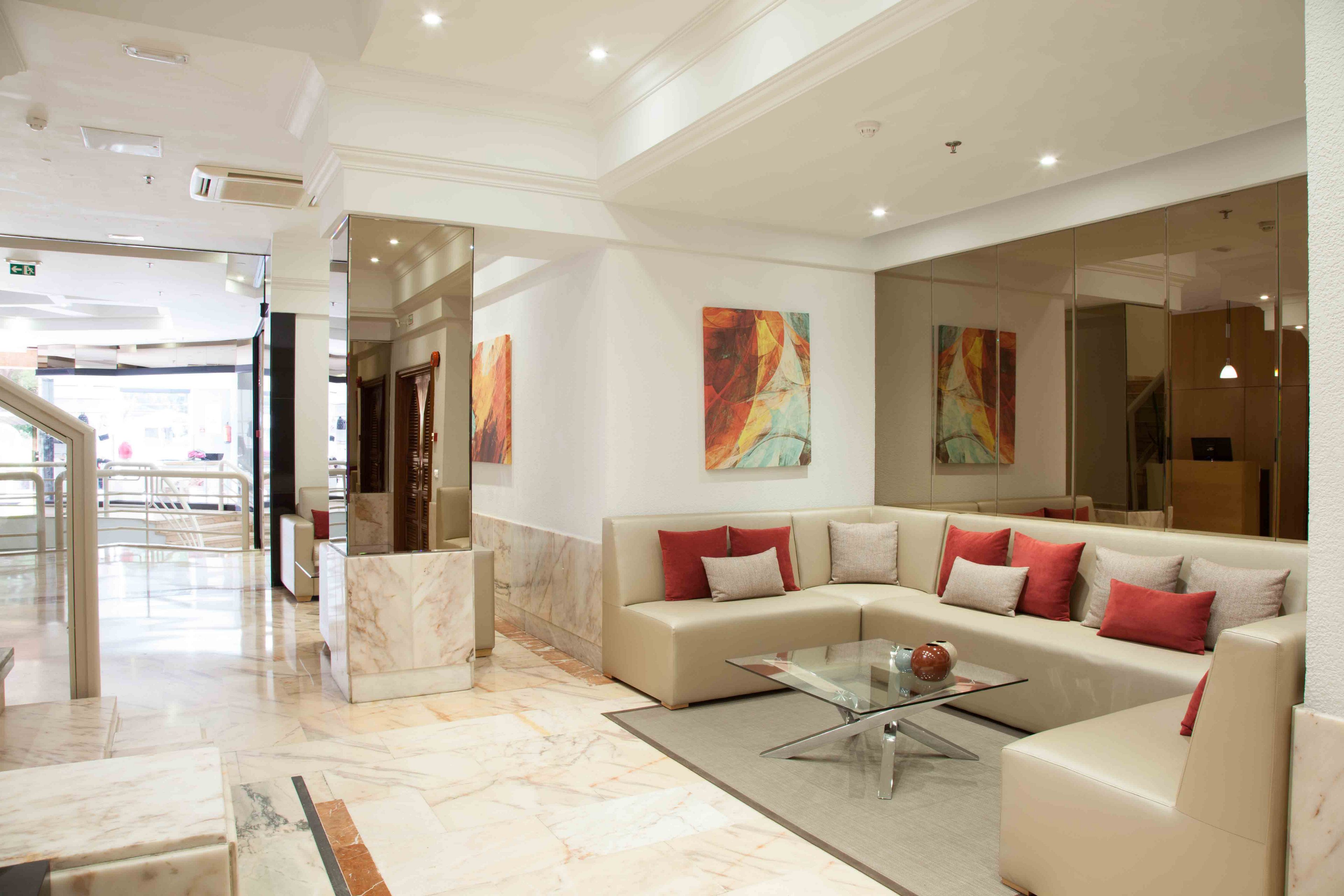 Bright contemporary lobby interior featuring a beige sectional sofa with red and white cushions. A glass coffee table sits in the center on a polished marble floor, with a decorative sphere placed on top. Abstract artwork with warm tones hangs on the walls, complementing the neutral color palette. Recessed ceiling lights and reflective surfaces create a clean, upscale atmosphere in the lounge area.