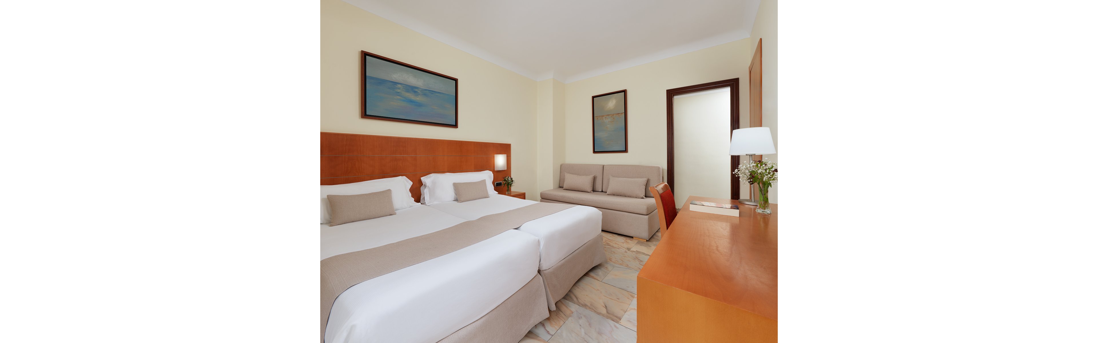 A bright, modern hotel room features two neatly made twin beds with white linens and beige accents. A beige sofa sits against the wall, complementing the light-colored walls and marble floor. Wooden furniture, including a desk and headboard, adds warmth to the space, while framed artwork and a table lamp provide decorative touches. A small vase with flowers on the desk enhances the clean, welcoming atmosphere.