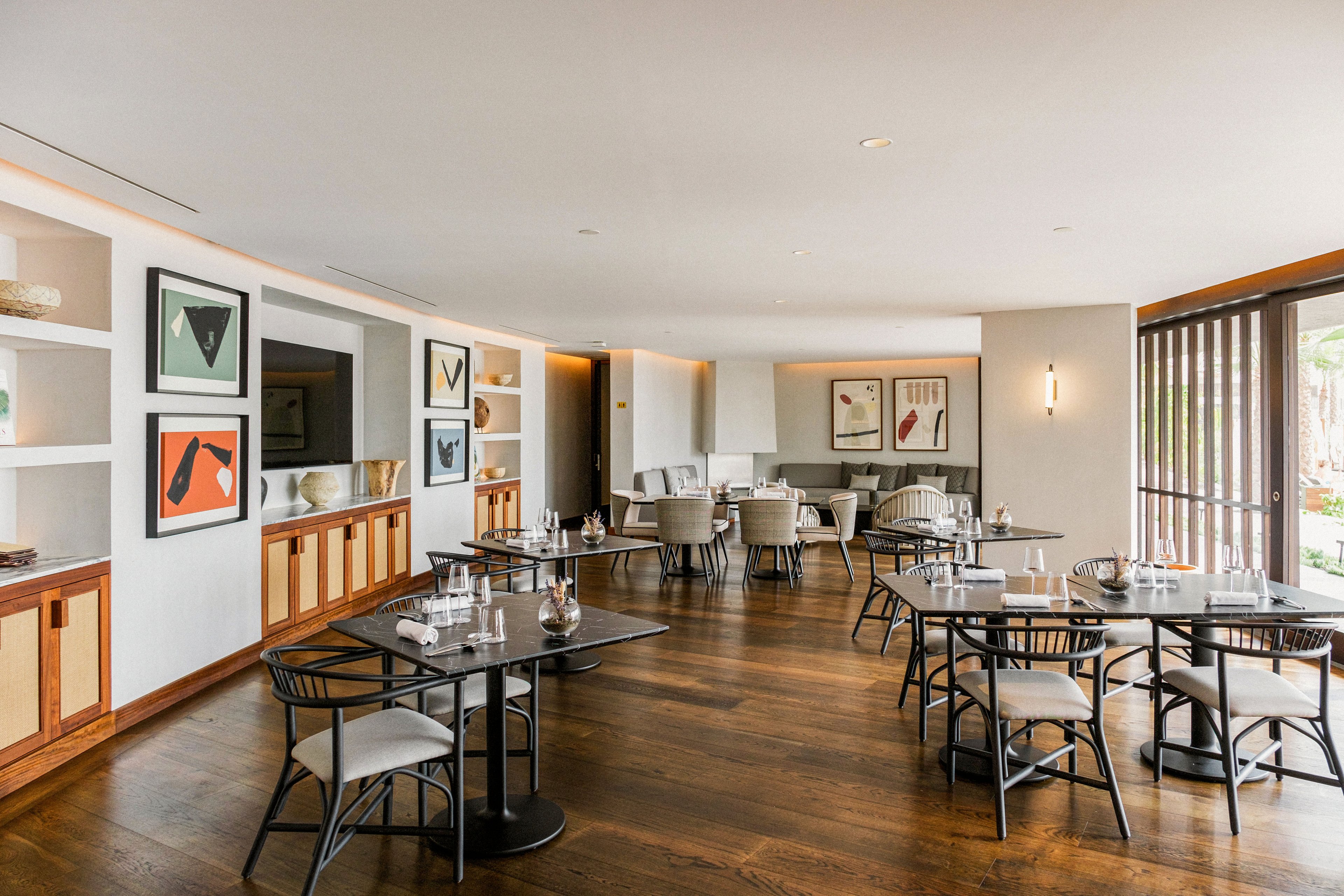 A contemporary restaurant dining area features neatly arranged tables and chairs on polished wooden flooring. Large windows on the right bring in natural light, highlighting neutral-toned furniture and minimalist decor. Framed abstract artwork adorns the walls, adding color and visual interest to the clean, modern interior. The space appears calm and inviting, suitable for relaxed dining or small gatherings.