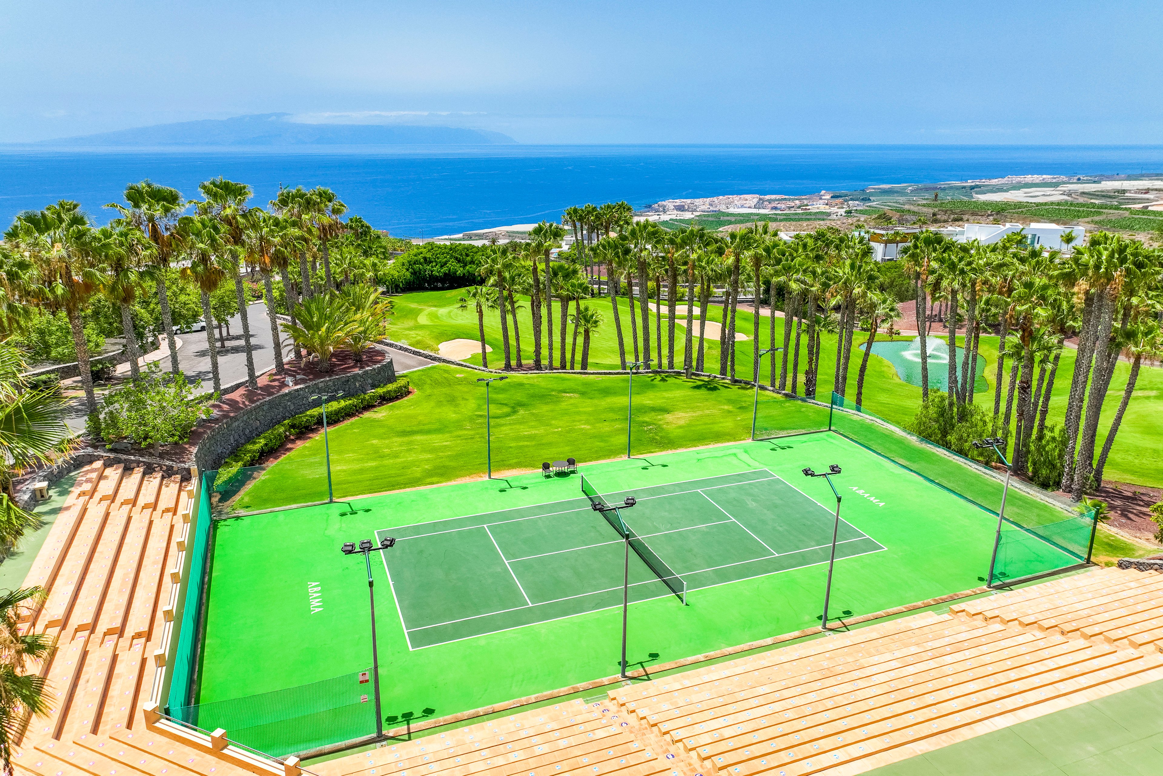 An elevated view shows a bright green tennis court framed by tall palm trees. The court is part of a landscaped resort area with terraced seating and manicured lawns. Beyond the sports facilities, the scene opens to a vivid blue ocean and distant island on the horizon. Clear skies and vibrant colors create a sunny, tropical atmosphere ideal for outdoor recreation.