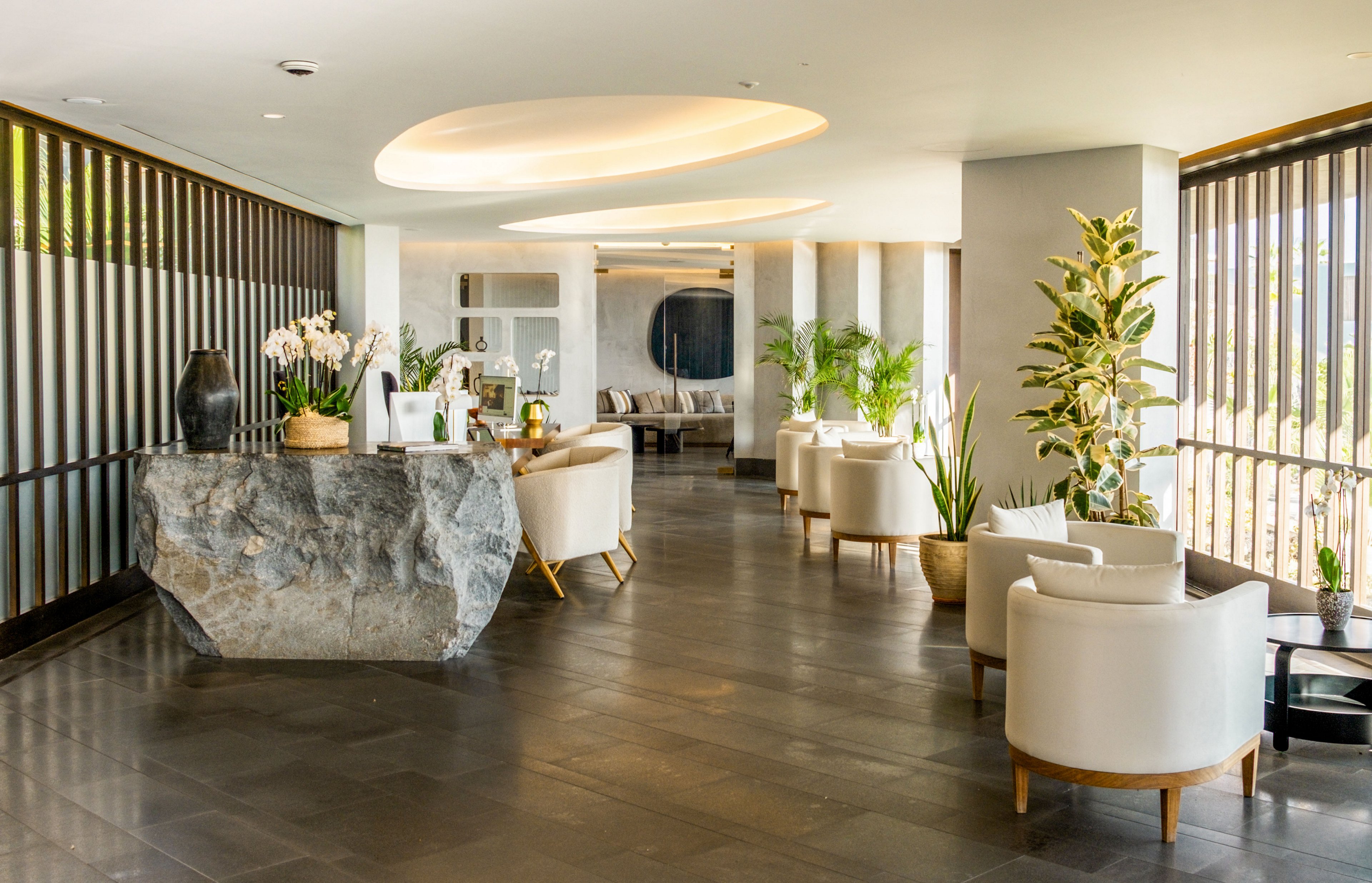 A contemporary hotel lobby features a sleek stone reception desk and comfortable white armchairs. Large floor-to-ceiling windows with vertical slats allow abundant natural light into the space. Potted plants and orchids add greenery and a touch of luxury to the minimalist interior. Warm ceiling lights and neutral tones create a calm, welcoming atmosphere.