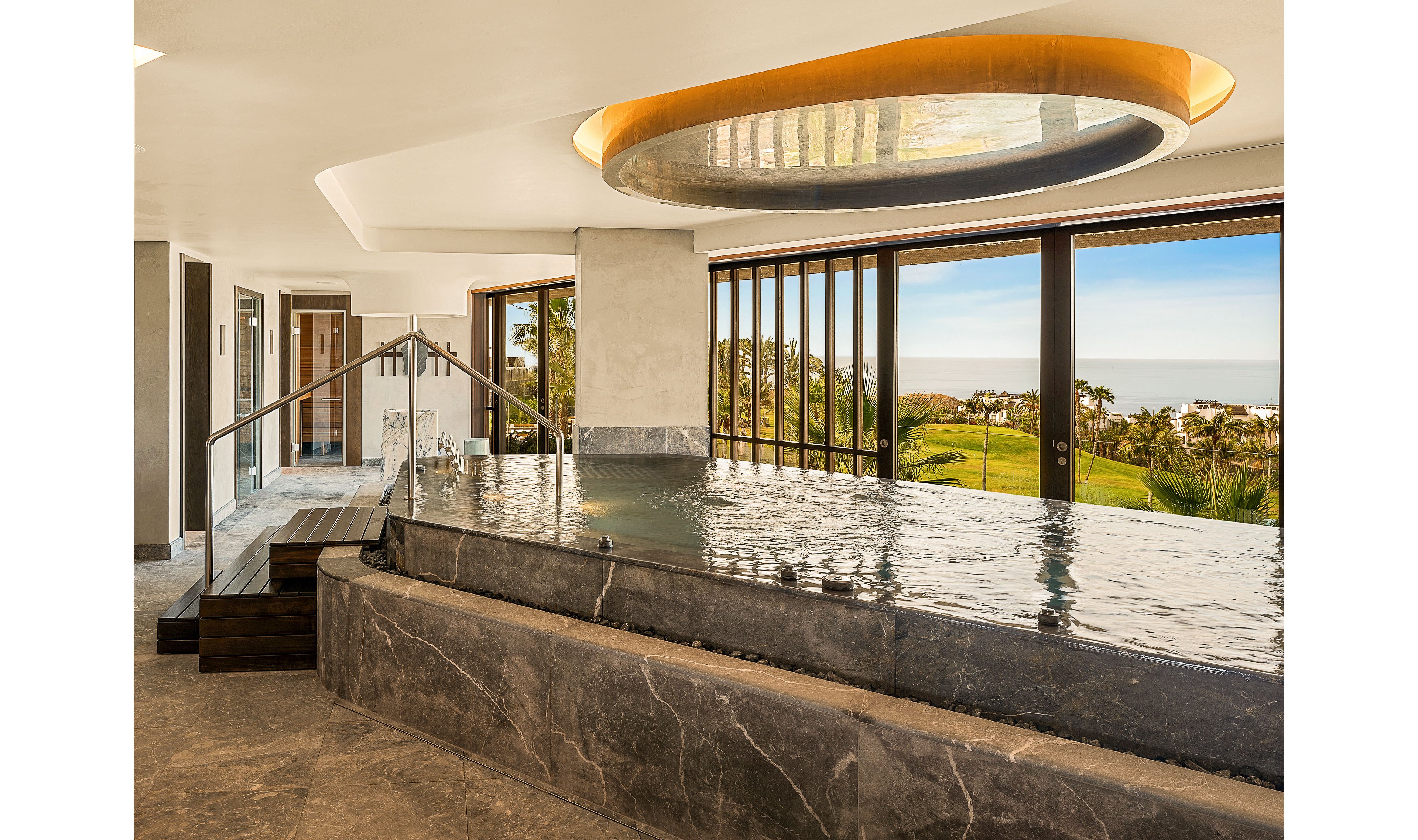 A sleek indoor infinity pool is featured in a contemporary spa-like setting. Large floor-to-ceiling windows reveal a lush green landscape and distant ocean horizon. The pool is framed by polished stone surfaces and a modern circular ceiling light above. Warm natural light fills the space, creating a tranquil and luxurious atmosphere.