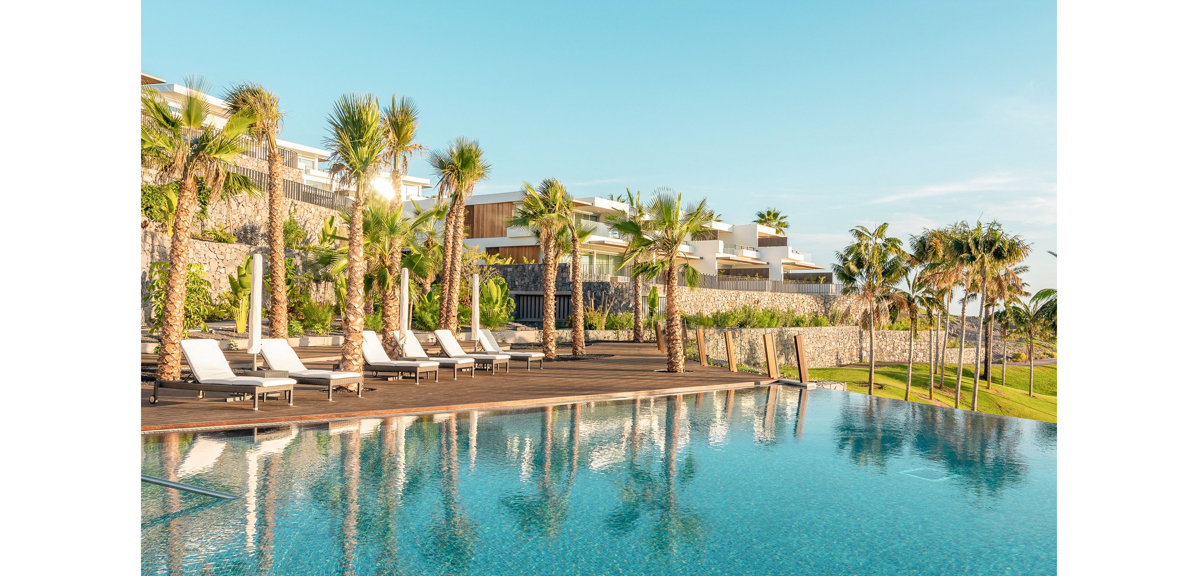 A luxurious resort pool stretches across the foreground, reflecting clear blue water and a bright sky. White lounge chairs line a wooden deck shaded by tall palm trees. Modern, low-rise buildings with stone walls and terraces rise behind the pool. The scene suggests a tranquil, tropical vacation setting with warm, golden morning light.