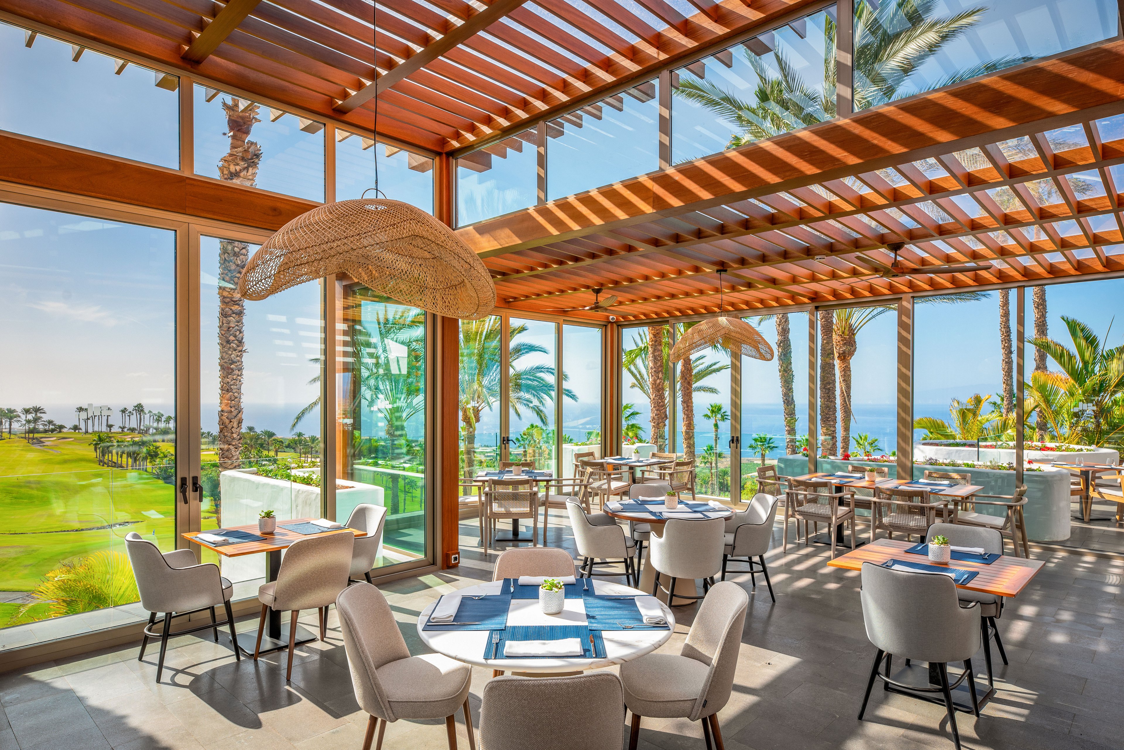 A modern, sunlit restaurant features neatly arranged tables and cushioned chairs facing large floor-to-ceiling windows. The space overlooks a lush green landscape with palm trees and a distant ocean view. Wooden ceiling slats and woven pendant lights add warmth and texture to the airy interior. Place settings and menus are prepared on each table, suggesting readiness for guests in a relaxed coastal dining atmosphere.