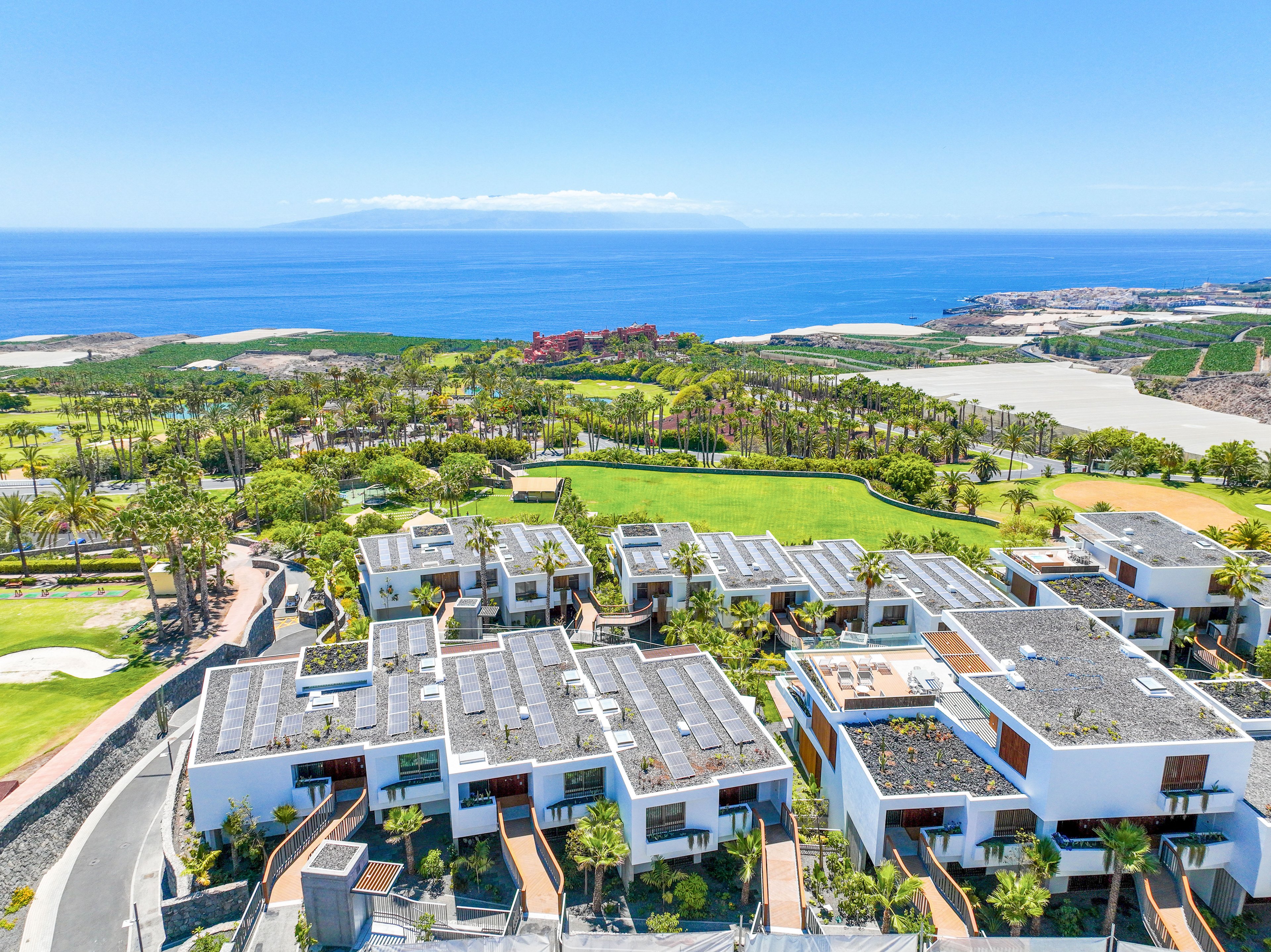 Aerial view of a modern coastal resort featuring white, flat-roofed buildings with rooftop terraces and solar panels. The complex is surrounded by lush greenery and palm trees, with a manicured golf course stretching toward the shoreline. In the distance, the blue ocean and a distant island create a scenic horizon under a clear sky. The image highlights contemporary architecture, sustainable design elements, and a luxurious seaside setting.