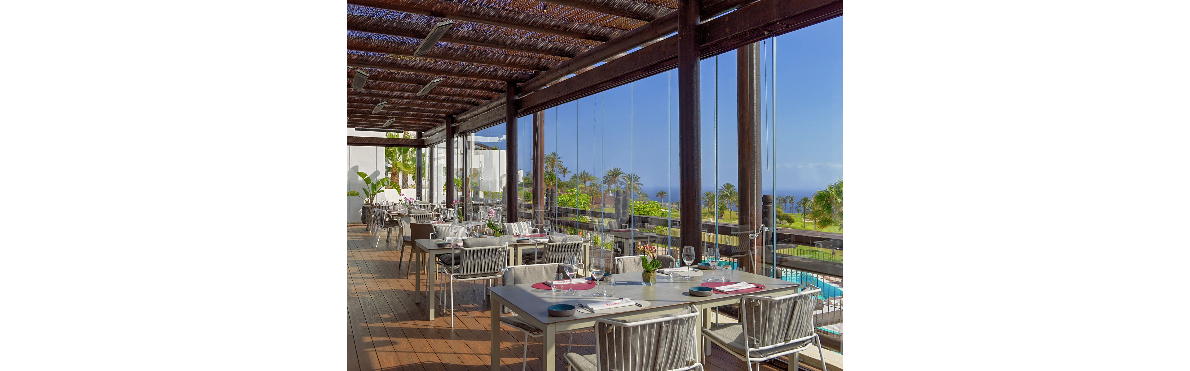 An elegant open-air restaurant terrace is set with neatly arranged tables and chairs. Large floor-to-ceiling glass panels reveal a view of a swimming pool, palm trees, and distant ocean. Sunlight filters through the wooden pergola ceiling, creating a warm and inviting atmosphere. Table settings include plates, glasses, and small floral centerpieces, suggesting a relaxed yet upscale dining experience.