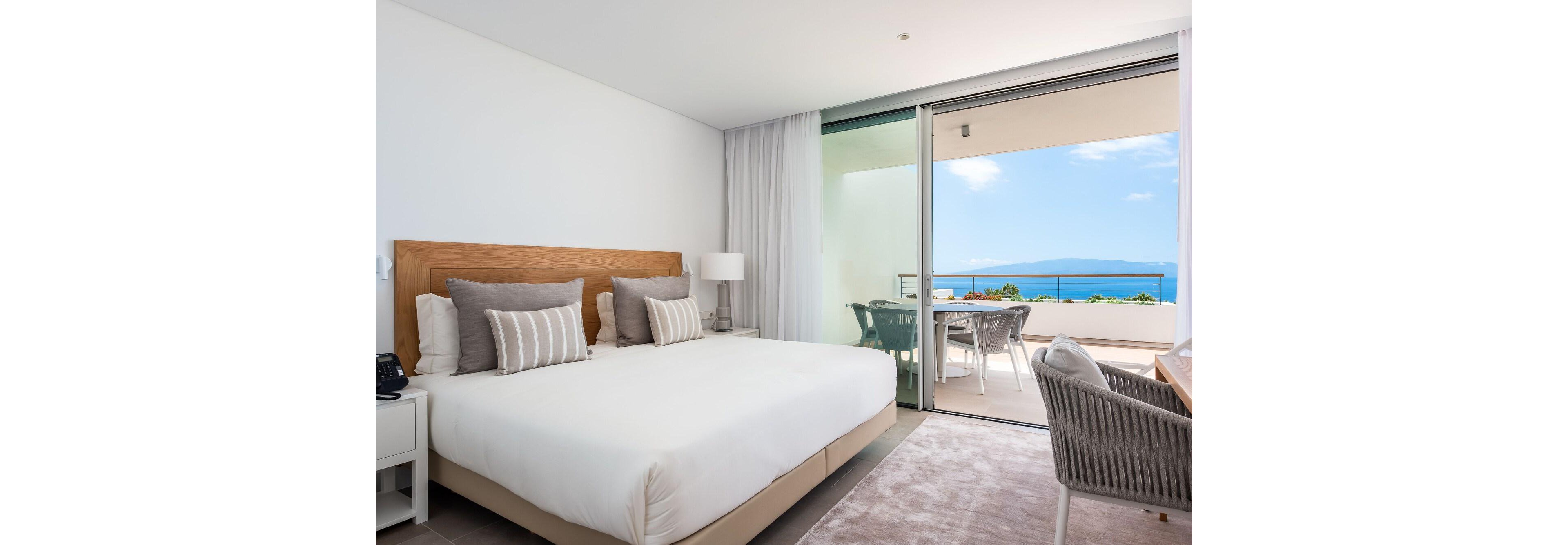 A bright, modern hotel bedroom features a large bed with white linens and striped accent pillows. The room opens onto a spacious balcony through sliding glass doors, revealing a clear view of the ocean and distant mountains. Neutral tones, soft textiles, and minimalist furniture create a calm, contemporary atmosphere. An outdoor dining table and chairs on the balcony invite guests to relax and enjoy the coastal scenery.