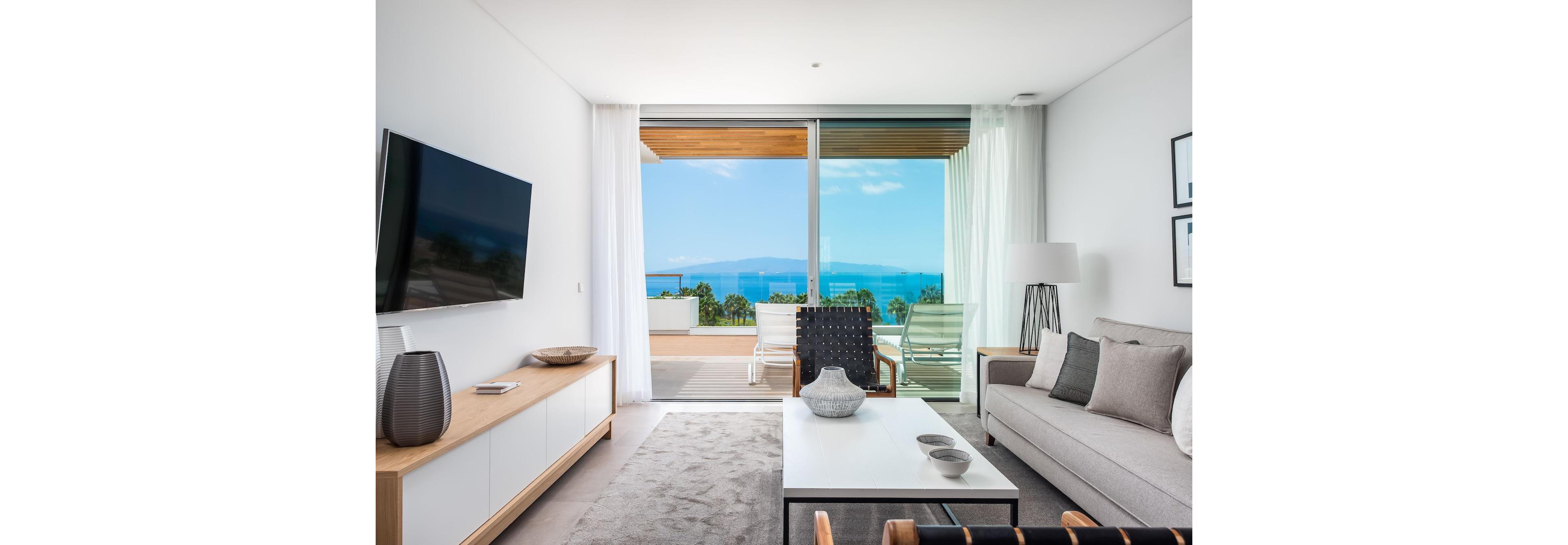 A contemporary living room features a sleek sofa, minimalist coffee table, and wall-mounted television. Large glass doors open onto a balcony with lounge chairs overlooking a bright blue ocean and distant mountains. White walls, light wood furniture, and neutral textiles create a clean, airy atmosphere. Soft natural light fills the space, emphasizing the modern coastal interior design.