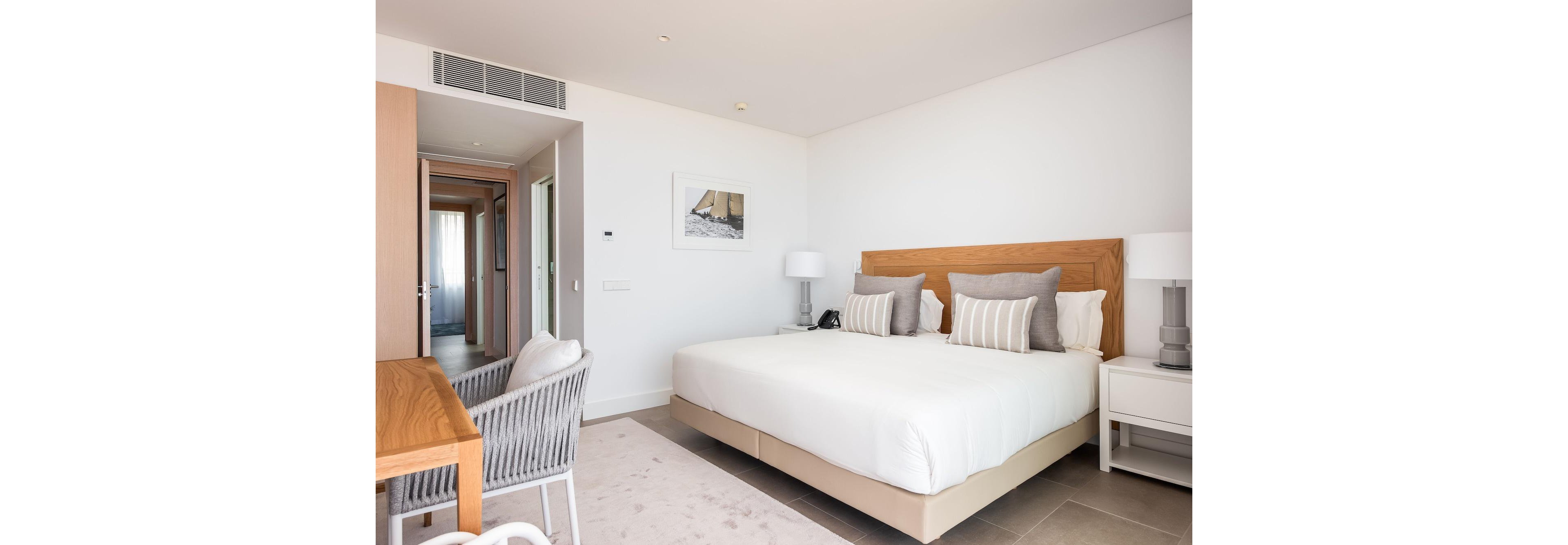 A contemporary hotel bedroom features a large bed with white linens and multiple gray and beige accent pillows. The room includes matching bedside tables with lamps, a desk with woven chairs, and a light wood headboard. Soft neutral tones dominate the space, complemented by a minimalist framed artwork on the wall. A hallway with natural light visible at the end adds depth and a sense of spaciousness to the interior.