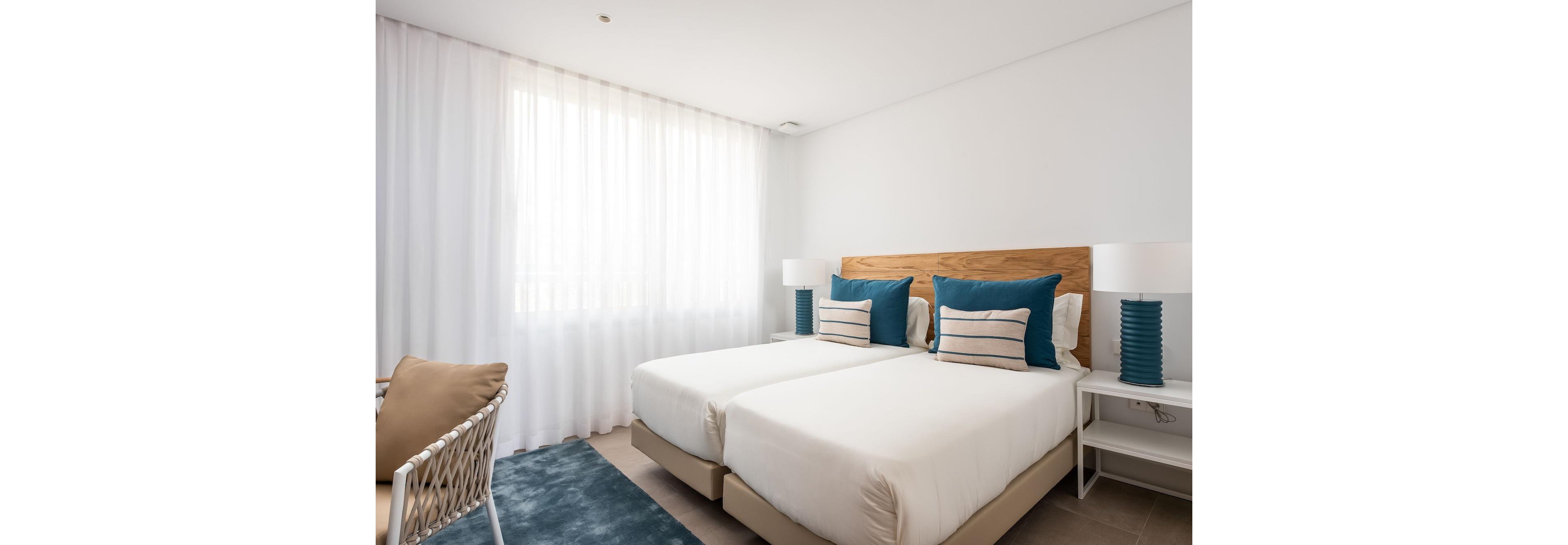 A bright modern bedroom features two neatly made twin beds with a shared wooden headboard. Teal pillows and striped cushions add color against the crisp white bedding and walls. Natural light filters through sheer white curtains, illuminating matching bedside tables with blue lamps. A woven chair and a blue area rug complete the cozy, minimalist decor.