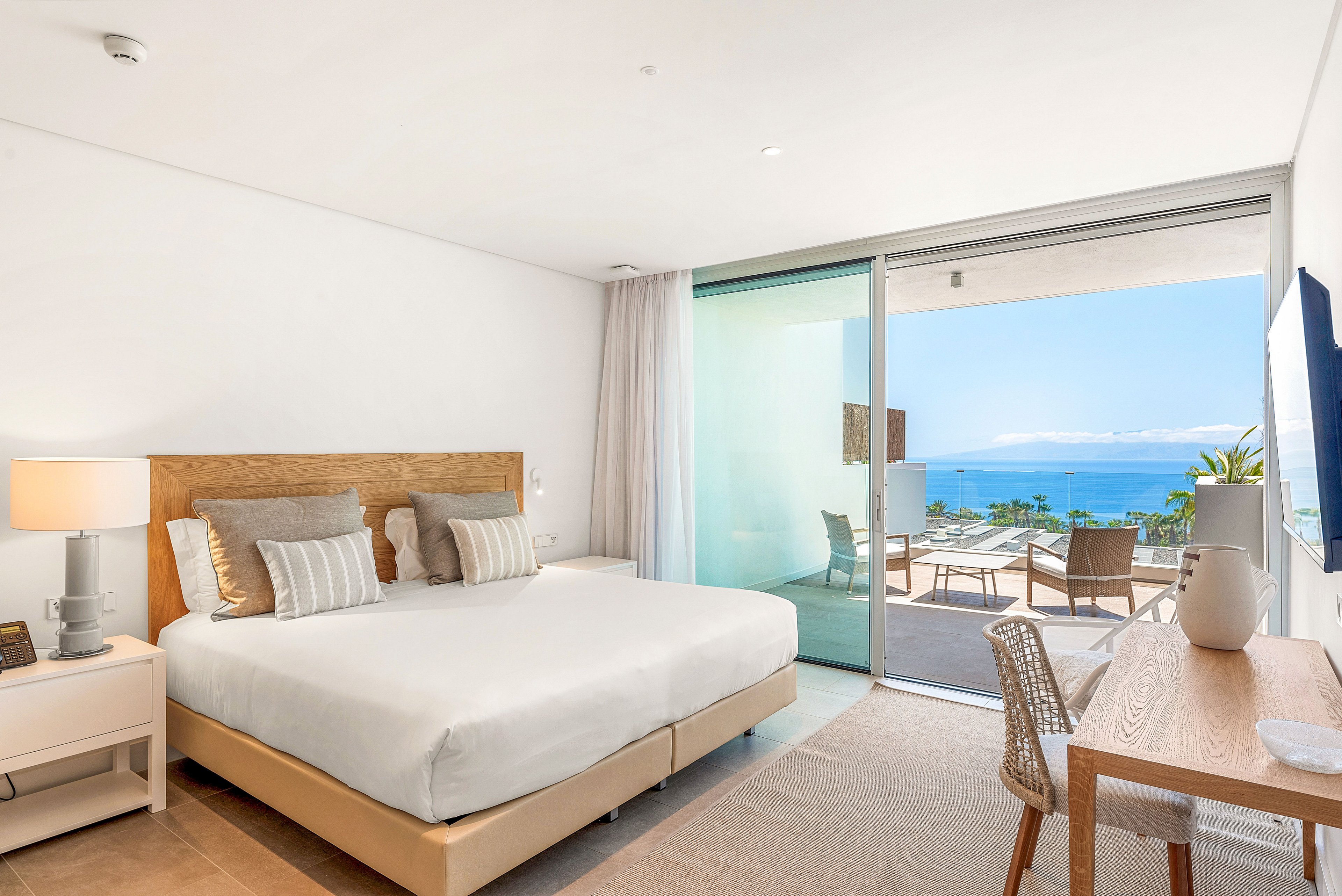 A bright, modern hotel bedroom features a large bed with neutral-toned pillows and linens. The room includes a wooden desk, chair, and bedside table with a lamp, all in a minimalist style. Large sliding glass doors open onto a terrace with lounge chairs and a table overlooking the ocean and distant coastline. Natural light fills the space, creating an airy and relaxing atmosphere ideal for a beachside vacation.