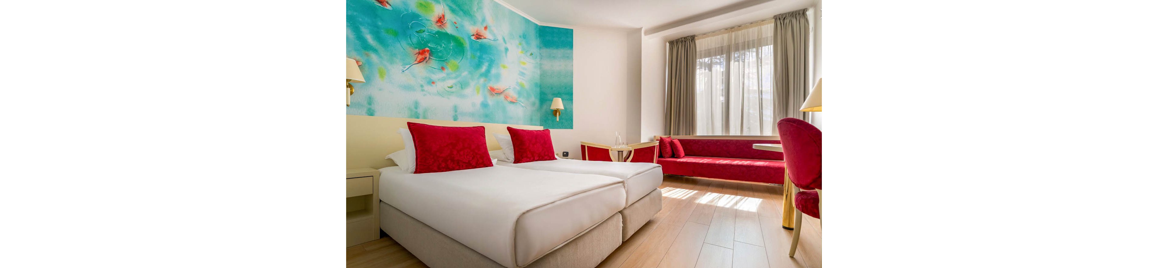 Contemporary hotel room featuring two neatly made twin beds with white linens and vivid red cushions. A long red upholstered bench runs along the window wall, adding extra seating and color. Large windows with sheer and heavy curtains allow natural light to fill the space. A turquoise wall mural with abstract fish motifs and small wall lamps creates a playful, artistic focal point.