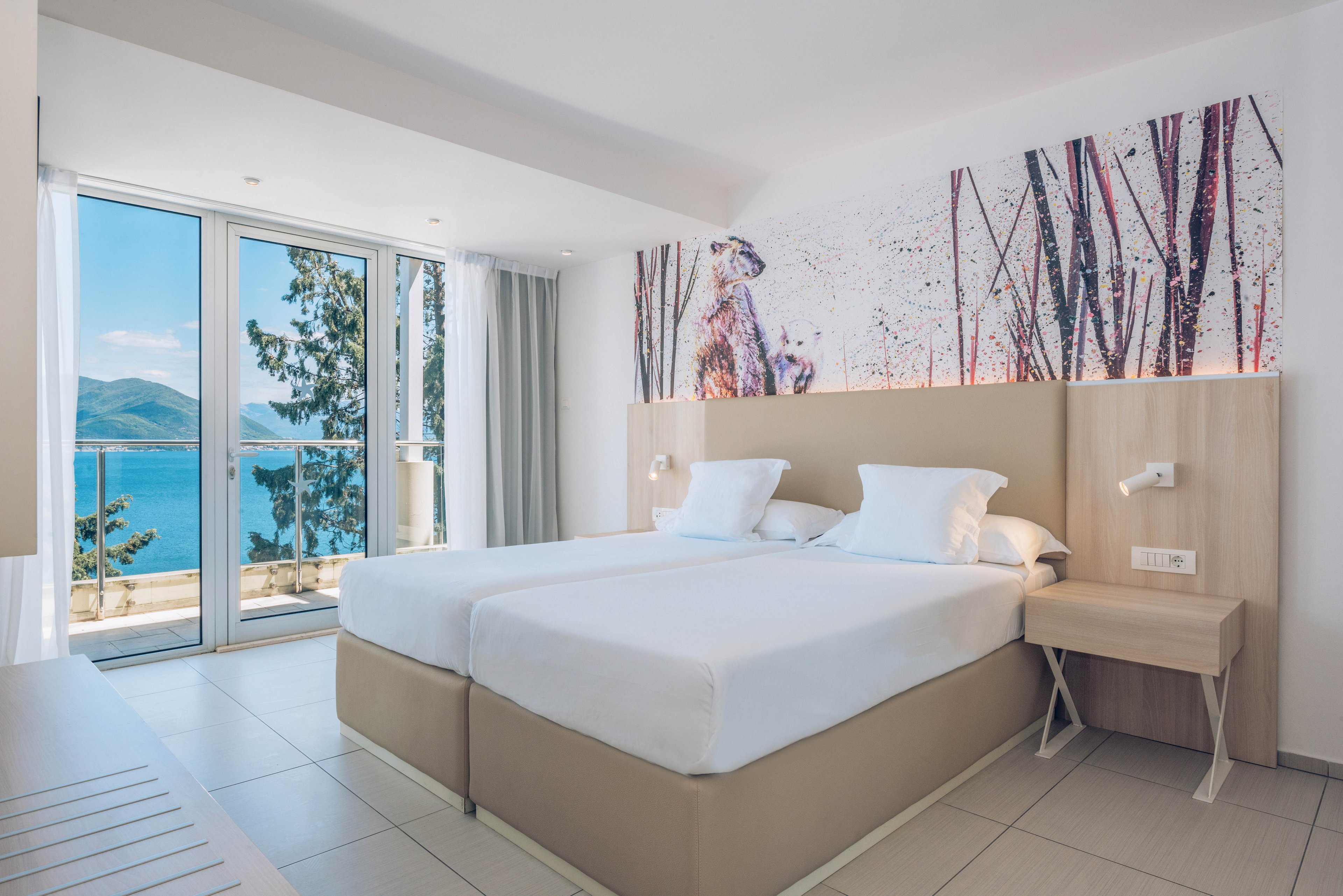 Bright modern hotel room featuring two neatly made twin beds with white linens and light wood furnishings. Large floor-to-ceiling glass doors open to a balcony with a scenic view of blue sea and distant mountains. A contemporary wall mural above the headboard adds artistic detail to the minimalist decor. Natural daylight fills the space, creating an airy and relaxing coastal atmosphere.