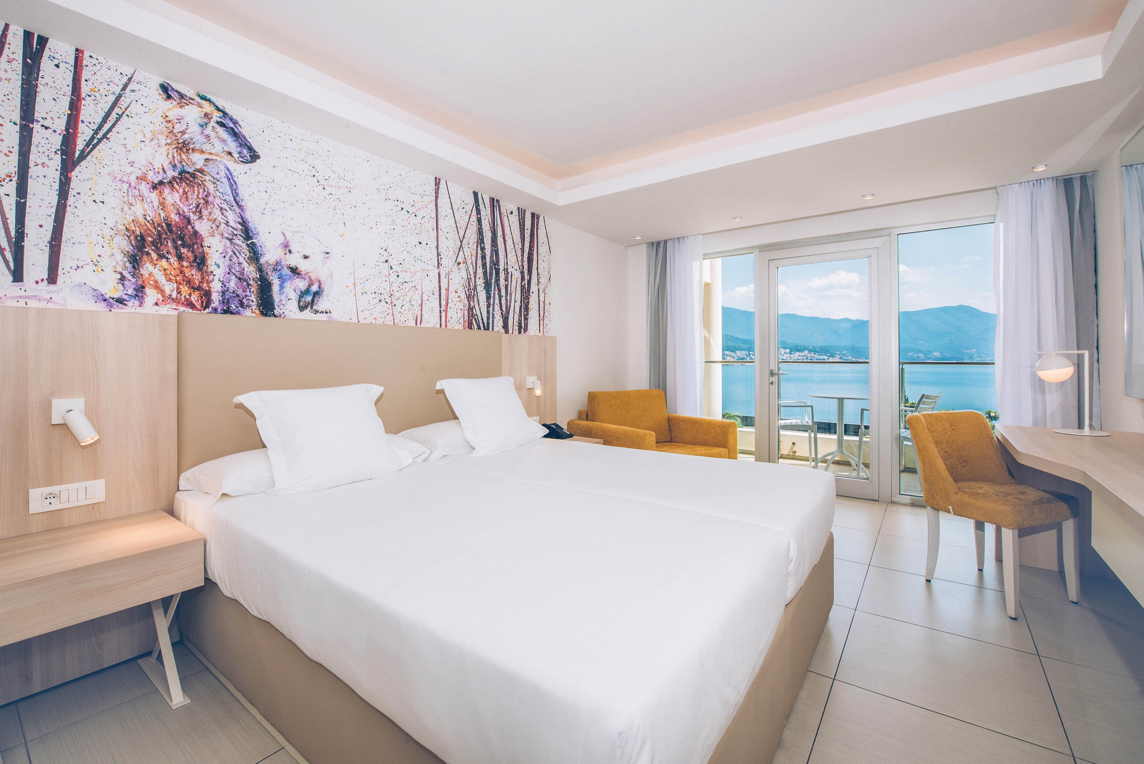 Bright modern hotel room featuring a large double bed with crisp white linens. A colorful wall mural of an animal adds artistic flair above the headboard. The room includes light wood furniture, a desk, and mustard-colored armchairs. Floor-to-ceiling windows open onto a balcony overlooking a calm sea and distant mountains, filling the space with natural light.