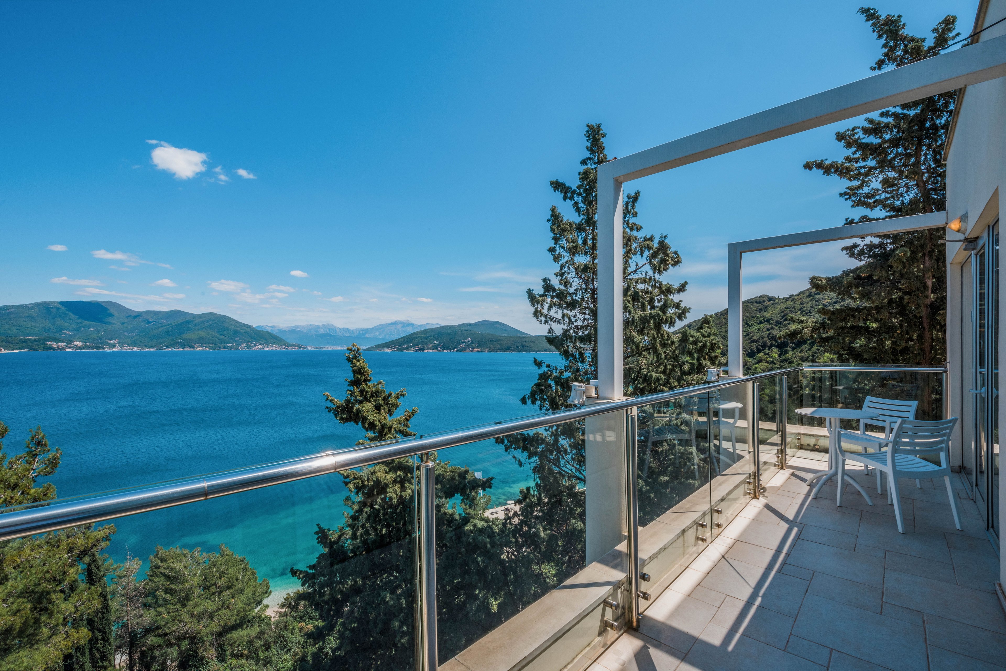 A modern balcony with glass railings overlooks a bright turquoise sea and distant green hills. The terrace features a small white table and chairs positioned to enjoy the coastal view. Tall evergreen trees rise from the slope below, framing the shoreline and water. Clear blue skies and strong sunlight create a serene, summery atmosphere.
