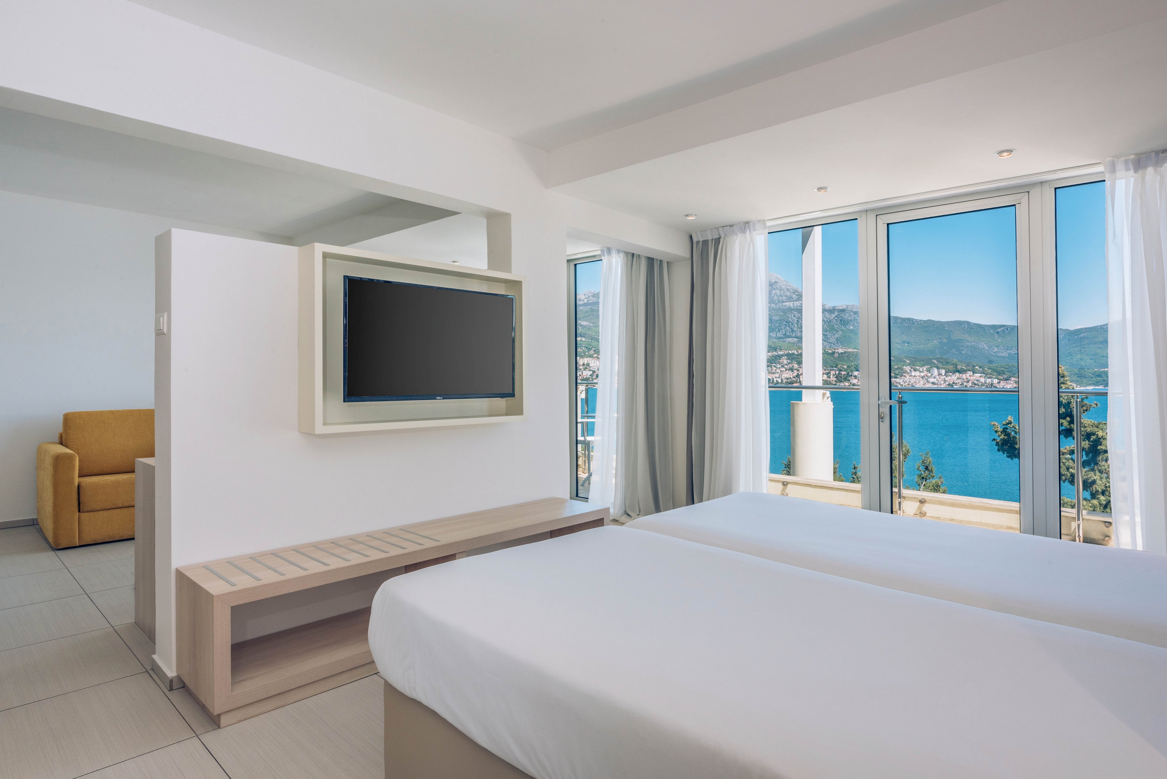 A bright, modern hotel room features two neatly made beds facing a wall-mounted flat-screen television. Large floor-to-ceiling windows open onto a stunning view of a blue sea and distant coastal town. Neutral white and beige tones dominate the interior, complemented by a mustard-colored armchair in the corner. Natural light floods the space, creating an airy and relaxing atmosphere ideal for a seaside vacation.