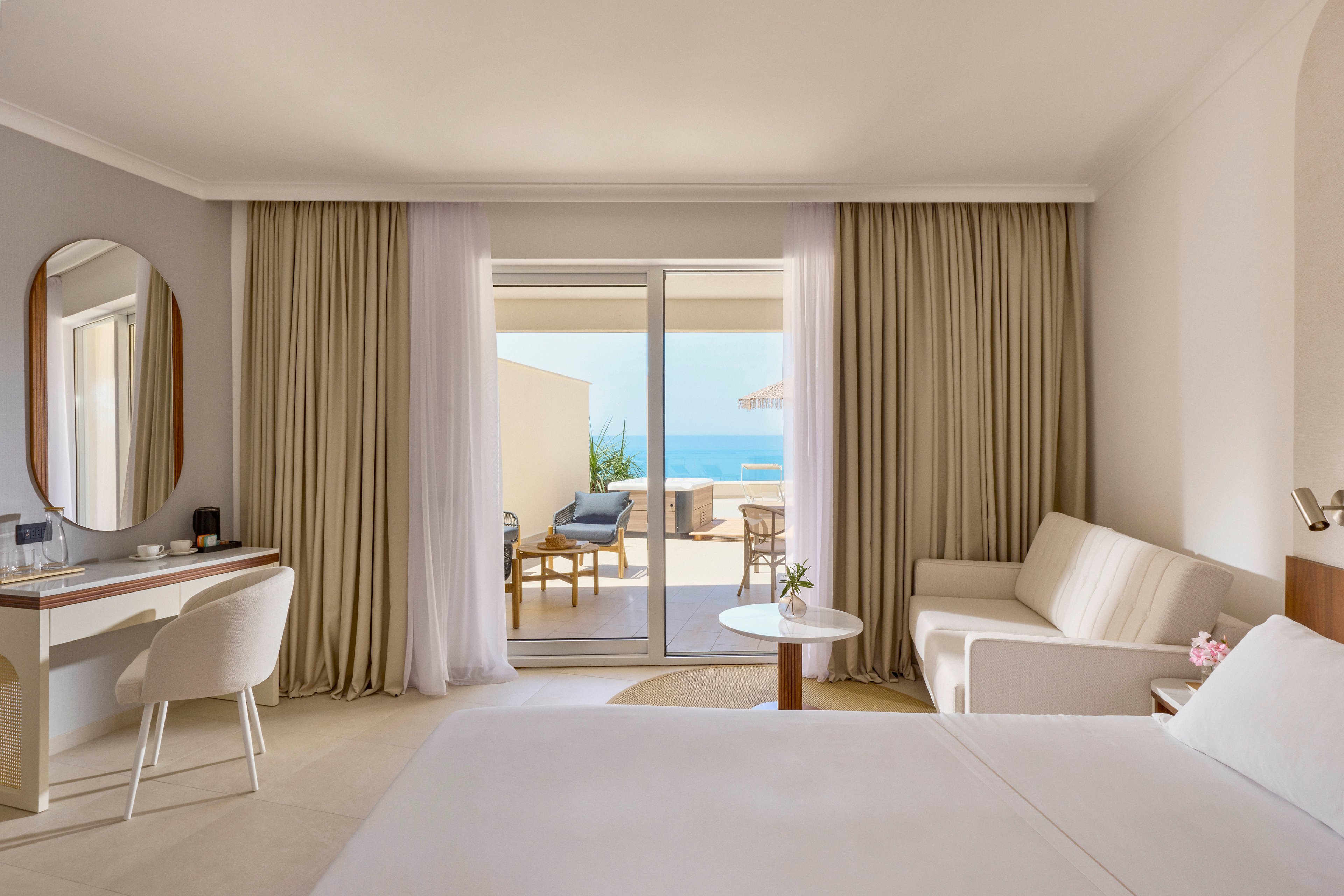 A contemporary hotel room features a neatly made bed, a white sofa, and a minimalist vanity desk with a round mirror. Floor-to-ceiling curtains frame sliding glass doors that open onto a private terrace with lounge chairs. Beyond the terrace, the blue ocean and clear sky create a serene coastal backdrop. Soft neutral tones and natural light give the space an airy, relaxing atmosphere.