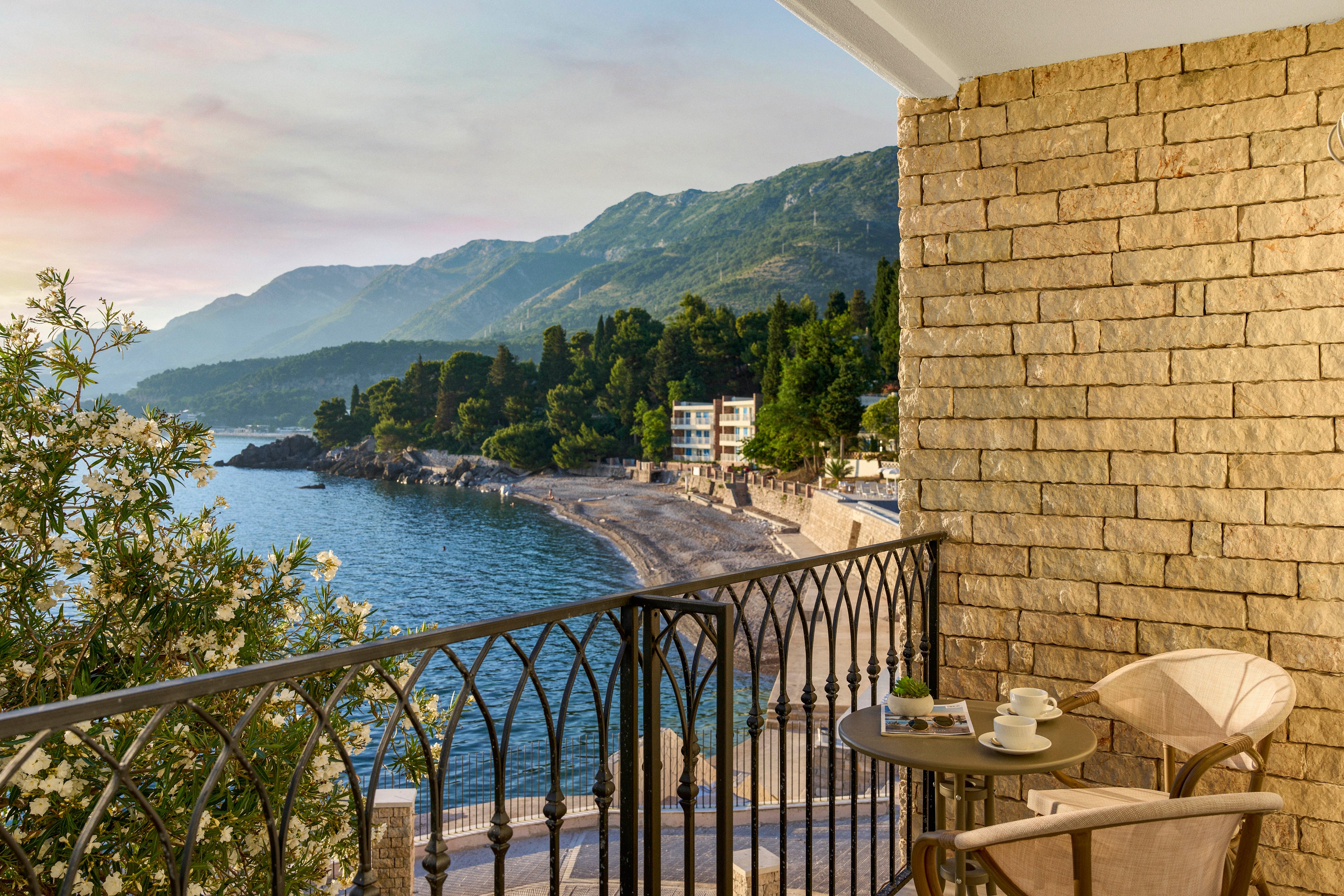 A cozy balcony overlooks a calm coastal bay with gentle waves and a distant shoreline. The scene includes a small round table set with drinks and a chair facing the water, framed by an ornate metal railing. Lush green hills and seaside buildings stretch along the coast under a soft, pastel sky at sunrise or sunset. Warm stone walls and flowering plants add texture and a relaxed Mediterranean ambiance.