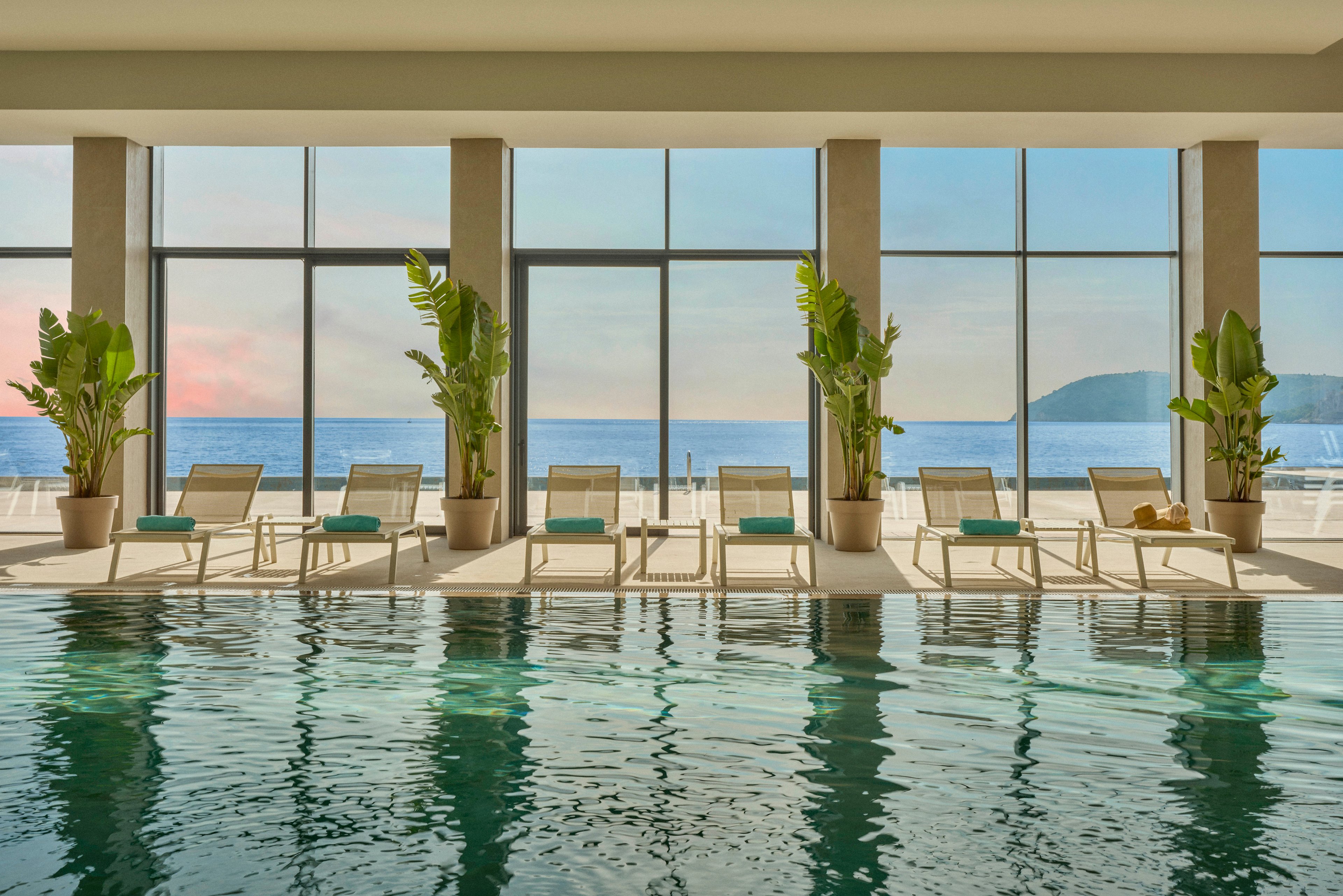 A serene indoor swimming pool is lined with modern lounge chairs and potted tropical plants. Floor-to-ceiling windows reveal a calm ocean and distant island at sunset, filling the space with soft natural light. The turquoise pool water reflects the chairs, plants, and sky, enhancing the tranquil atmosphere. The scene suggests a luxurious coastal resort or spa focused on relaxation and comfort.