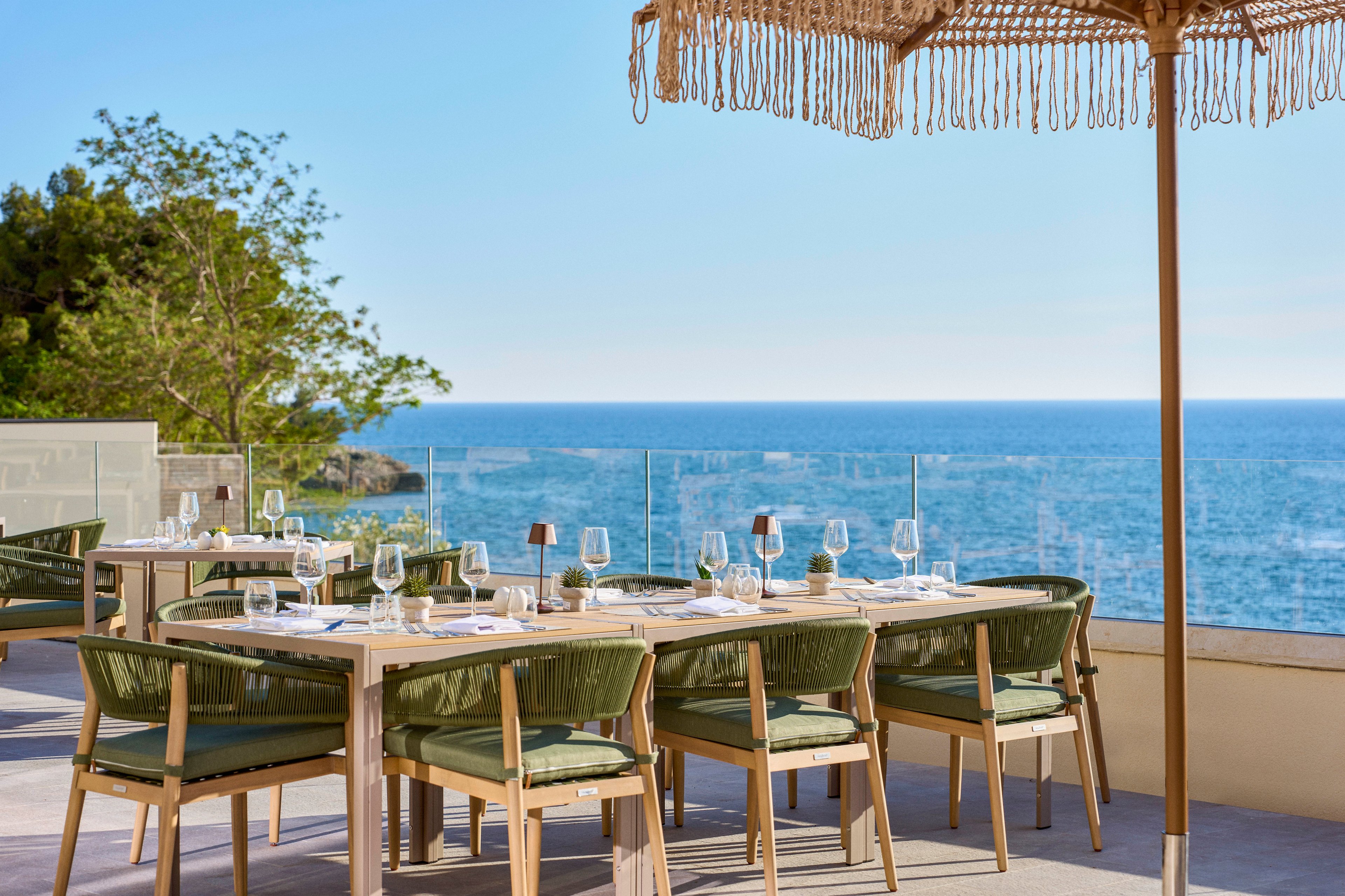 An elegant outdoor restaurant terrace overlooks a calm blue ocean under a clear sky. Wooden tables and woven chairs are neatly arranged with glassware, plates, and napkins ready for guests. A thatched parasol provides shade, while glass railings frame the scenic coastal view. Lush greenery on the left adds a natural touch to the relaxed, upscale dining atmosphere.