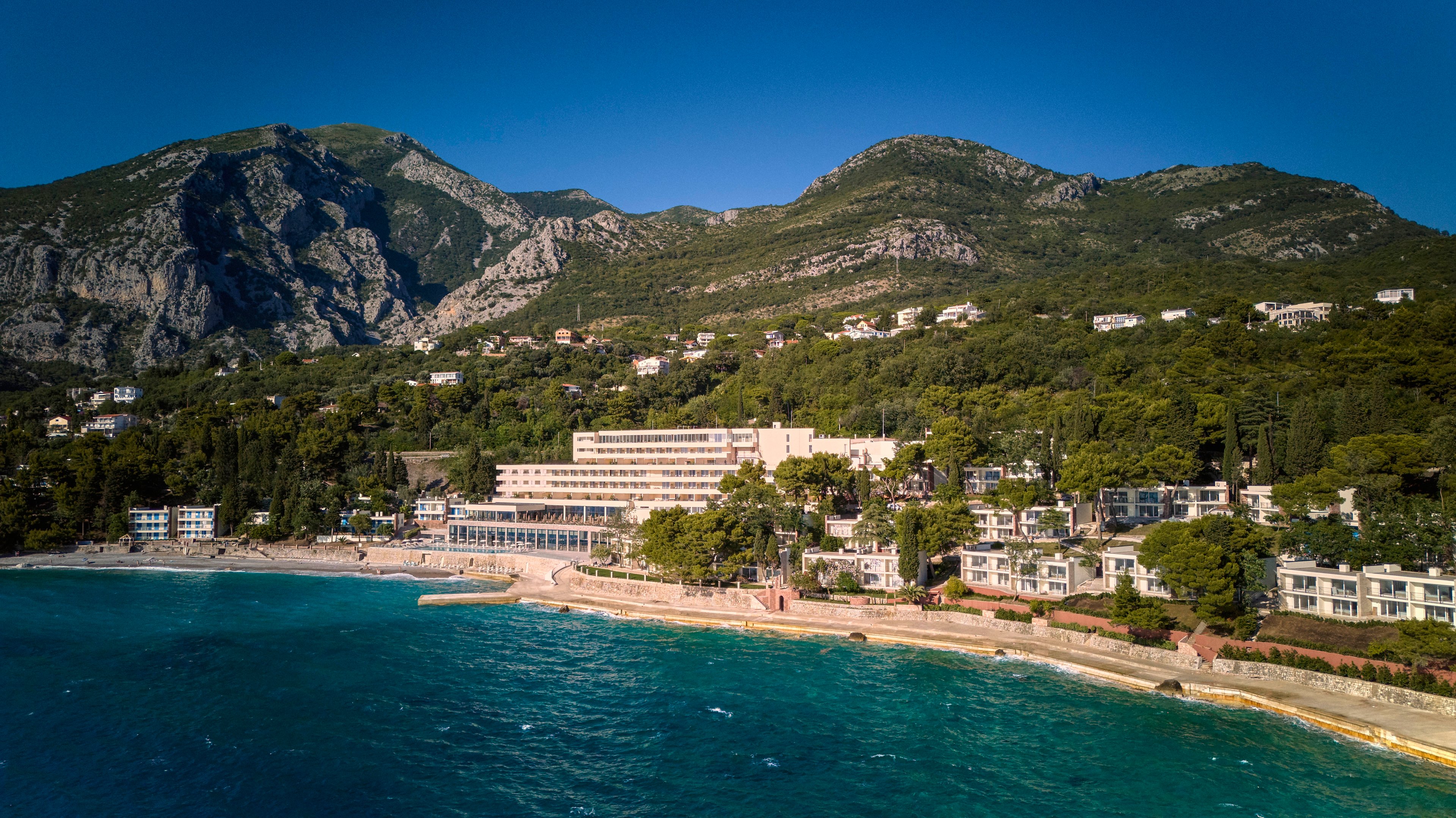 A seaside resort complex stretches along a turquoise shoreline at the base of a tall, rugged mountain range. White and pastel buildings line the coast, bordered by dense green forest and a narrow sandy beach. The calm blue sea occupies the foreground, while the mountains rise dramatically in the background under a clear sky. The scene suggests a tranquil vacation destination with natural beauty and coastal charm.