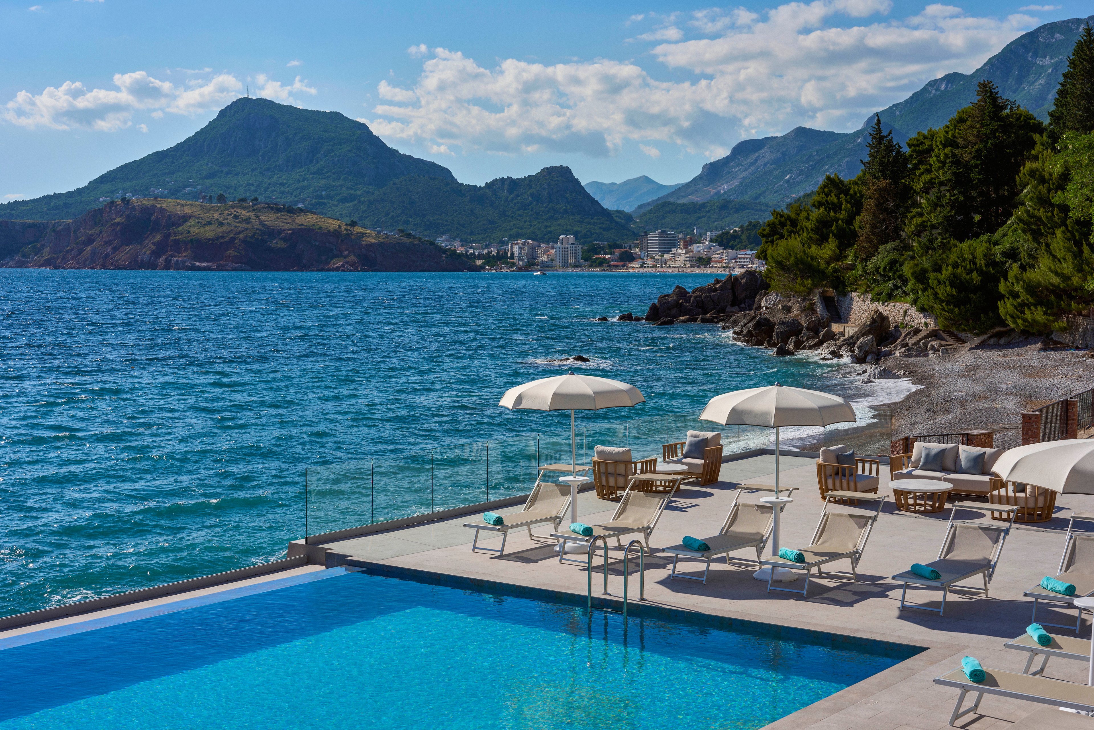 A luxurious infinity pool sits beside a rocky shoreline, facing a calm blue sea. White sun loungers and umbrellas line the pool deck, creating a relaxed resort atmosphere. Green hills and dramatic mountains rise in the background, with a coastal town visible along the distant bay. Clear skies with scattered clouds enhance the bright, tranquil vacation setting.