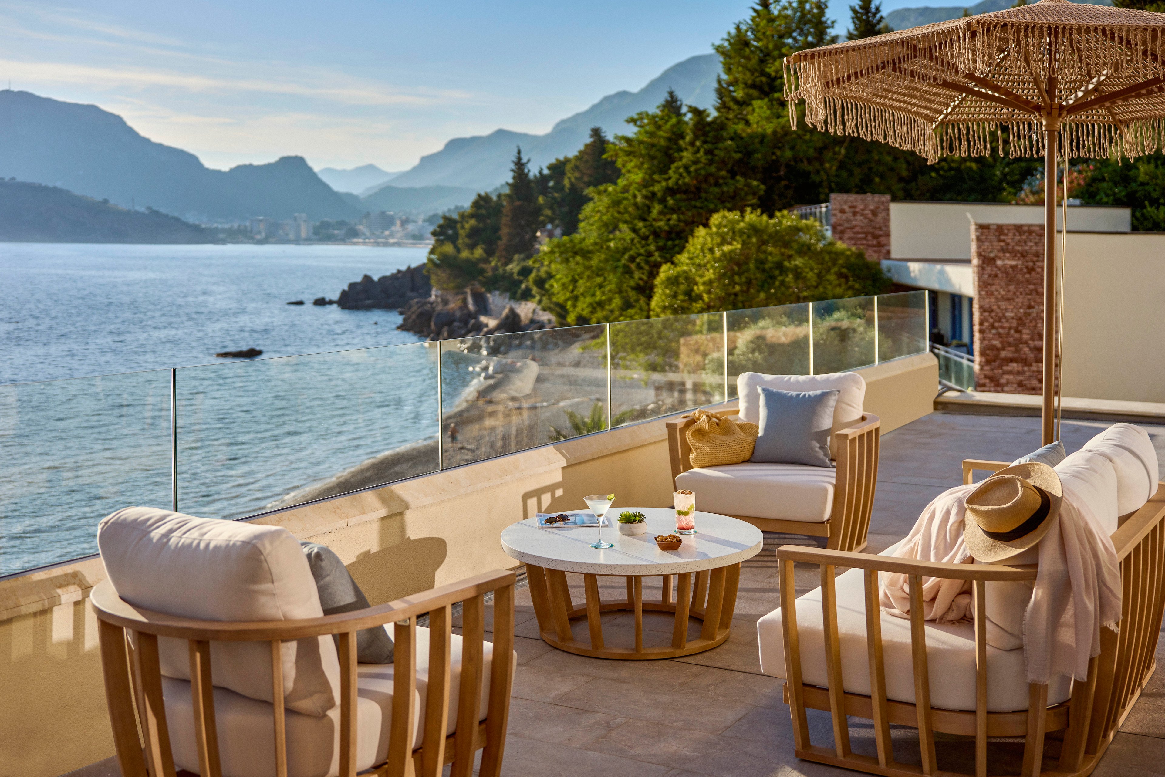 A stylish coastal terrace overlooks a calm sea and distant mountains at sunset or early evening. Comfortable cushioned lounge chairs surround a round white table with drinks and snacks, suggesting a relaxed vacation setting. A straw hat and light clothing draped on a chair imply a leisurely moment in warm weather. Glass railings, modern architecture, and a woven parasol add to the upscale, tranquil atmosphere.