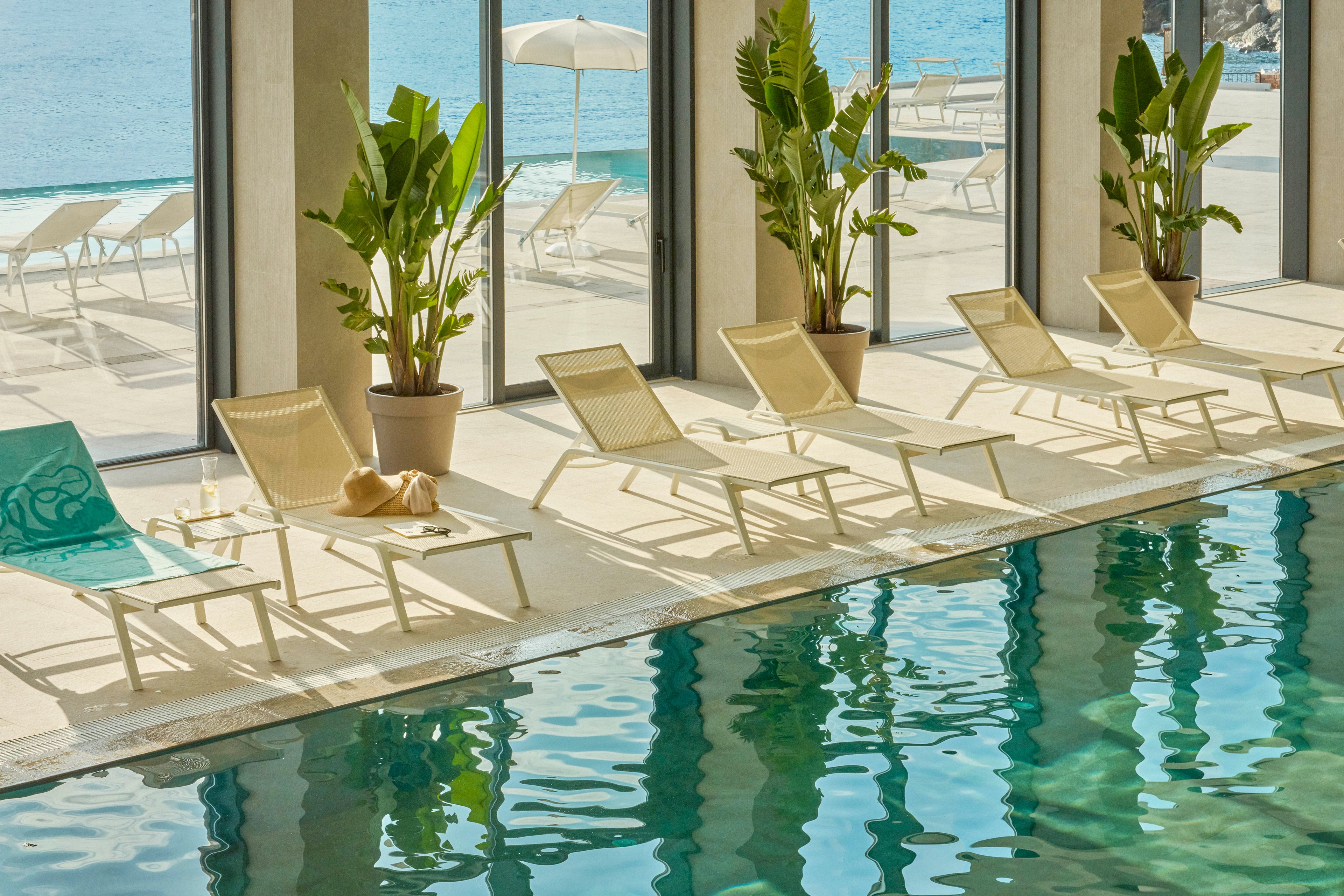 A serene indoor pool is lined with light-colored lounge chairs and potted tropical plants beside large floor-to-ceiling windows. Sunlight reflects off the turquoise water, creating shimmering patterns on the pool surface and surrounding tiles. Outside, a bright seaside terrace with additional loungers and umbrellas overlooks a calm blue ocean. The scene conveys a relaxed, resort-style atmosphere ideal for leisure and vacation.