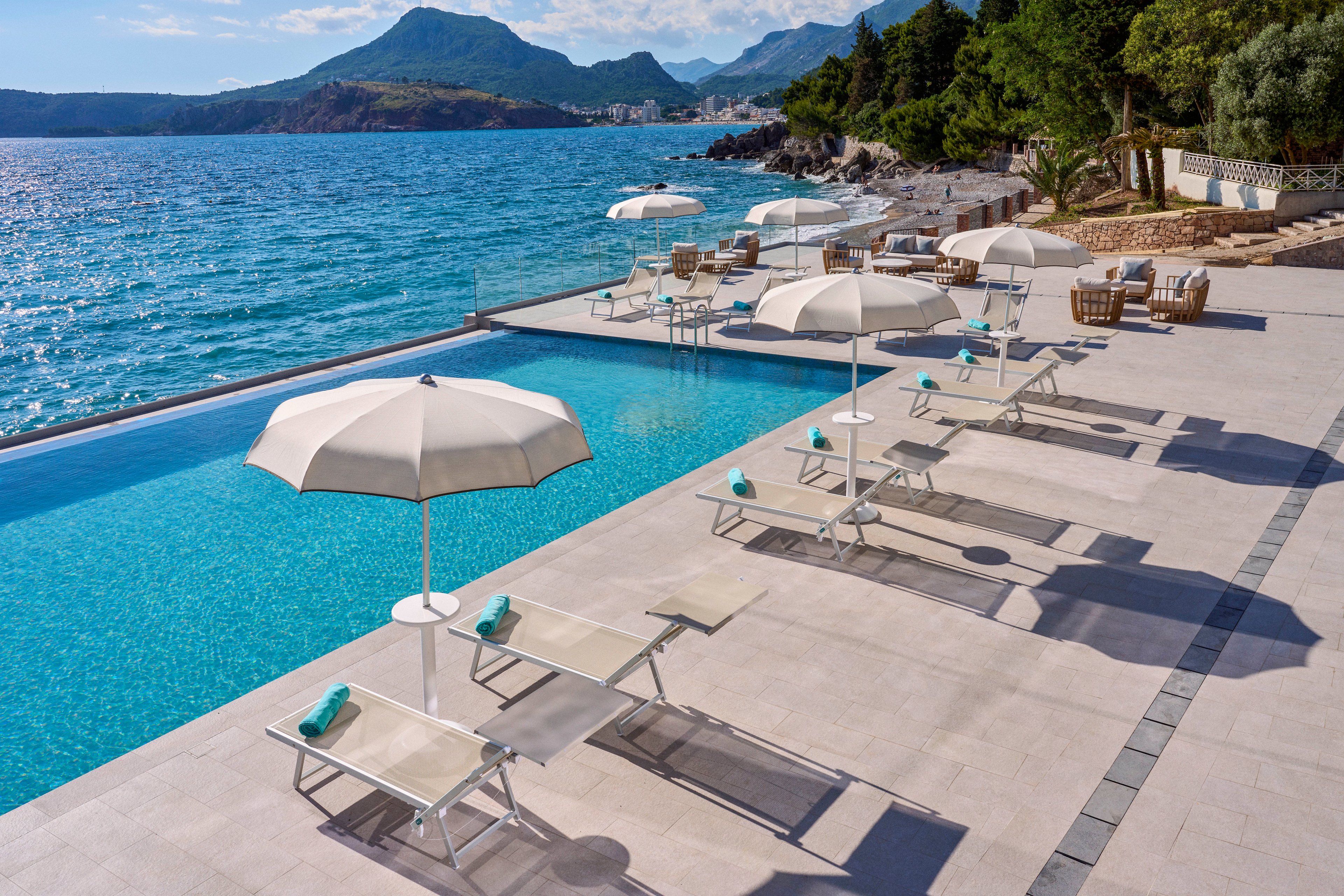 A serene infinity pool overlooks a calm blue sea with mountains in the distance. White sun loungers and umbrellas line the poolside terrace, each with a neatly rolled turquoise towel. The setting appears to be a high-end coastal resort, with additional outdoor seating areas visible in the background. Bright sunlight, clear skies, and clean modern design create a relaxing vacation atmosphere.