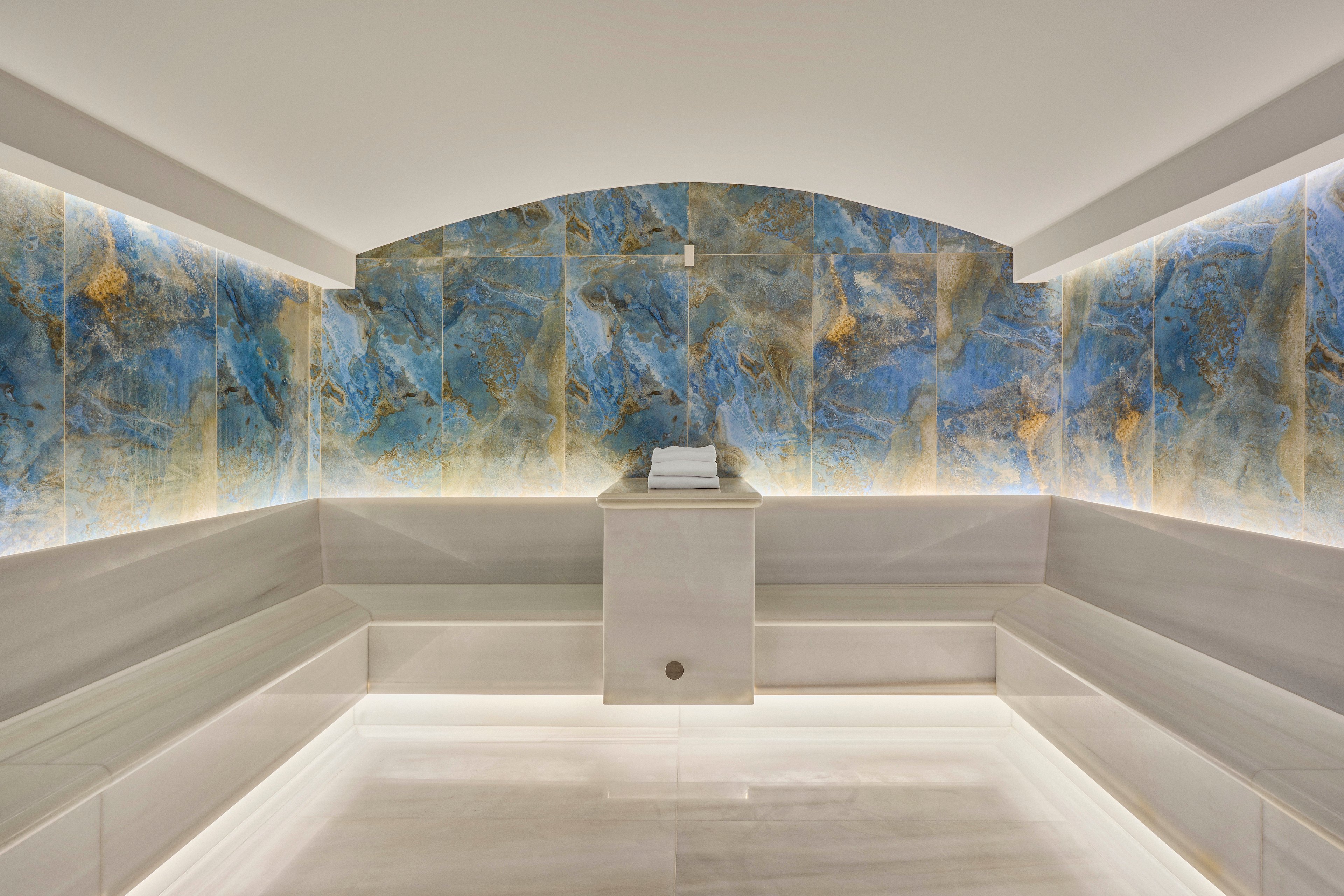 A contemporary spa steam room features sleek white benches and a central pedestal with neatly folded towels. The back wall is covered in striking blue and gold stone panels, softly illuminated by concealed lighting. Clean lines, neutral tones, and indirect light create a calm, luxurious atmosphere. The space appears empty, emphasizing tranquility and relaxation.