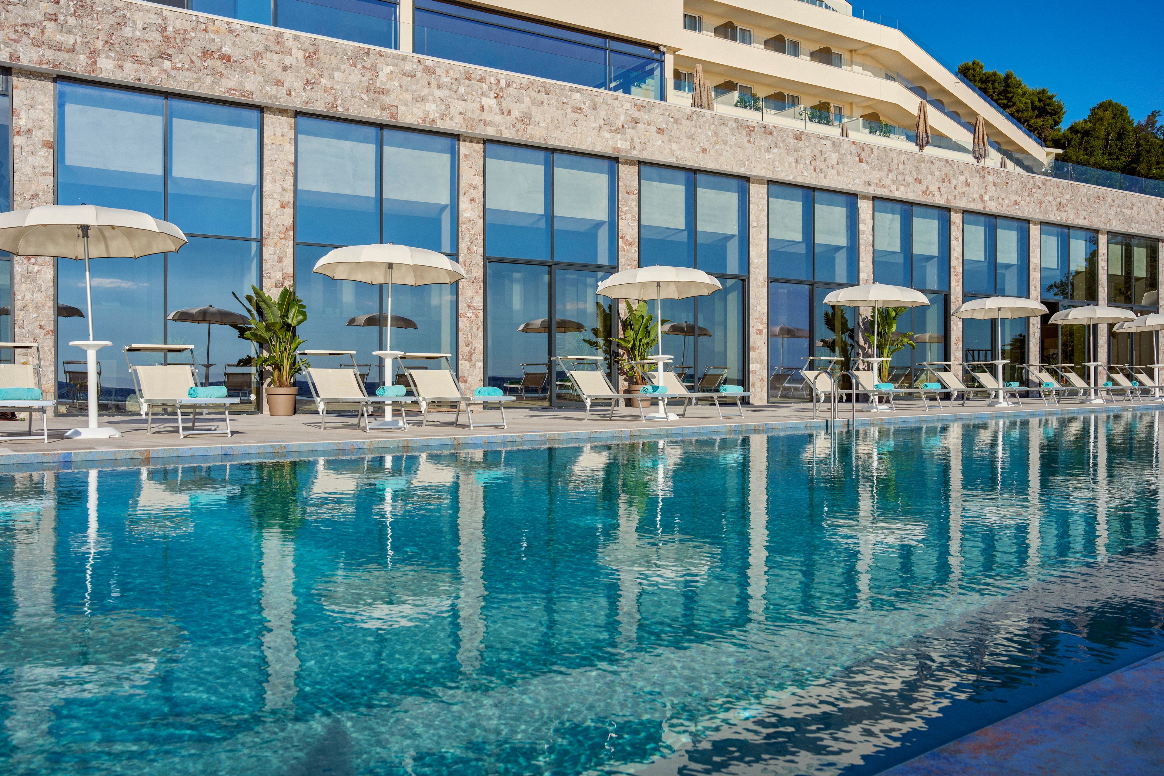 A large outdoor swimming pool reflects the modern glass façade of a resort building. White sun loungers and umbrellas line the poolside terrace, creating a clean and organized relaxation area. Potted tropical plants add greenery between the seating areas, while the stone and glass architecture gives a contemporary coastal feel. The scene suggests a sunny, tranquil vacation setting with clear blue water and bright skies.
