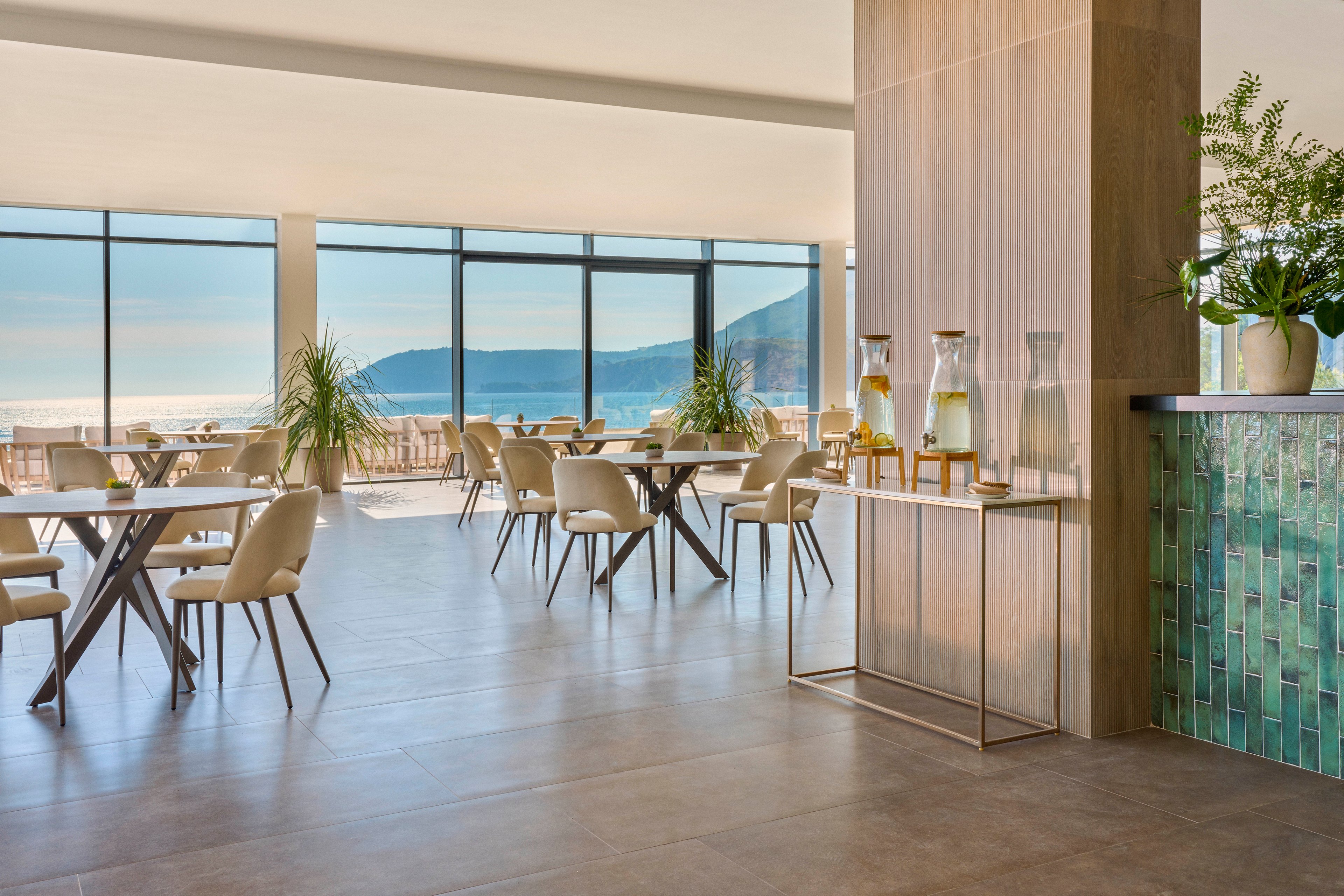 Bright contemporary dining area with large floor-to-ceiling windows overlooking the sea and distant hills. Minimalist tables and upholstered chairs are arranged neatly across the spacious room. A refreshment station with glass beverage dispensers and carafes stands beside a column, complemented by green tiled wall accents and potted plants. Natural light and neutral tones create an airy, relaxed coastal atmosphere.