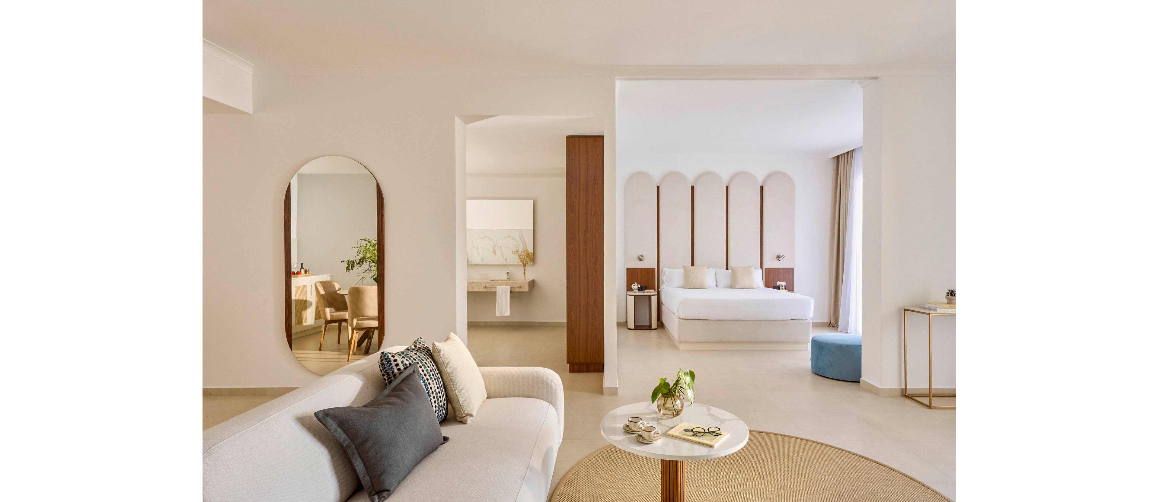 Bright contemporary suite features a white sofa and round coffee table in the foreground. The open-plan space leads to a bedroom with a large bed framed by arched wall panels and soft neutral textiles. A dining or work area with upholstered chairs is visible through an archway, complemented by a tall wall mirror. Light beige walls, natural wood accents, and subtle decor create a calm, minimalist atmosphere.