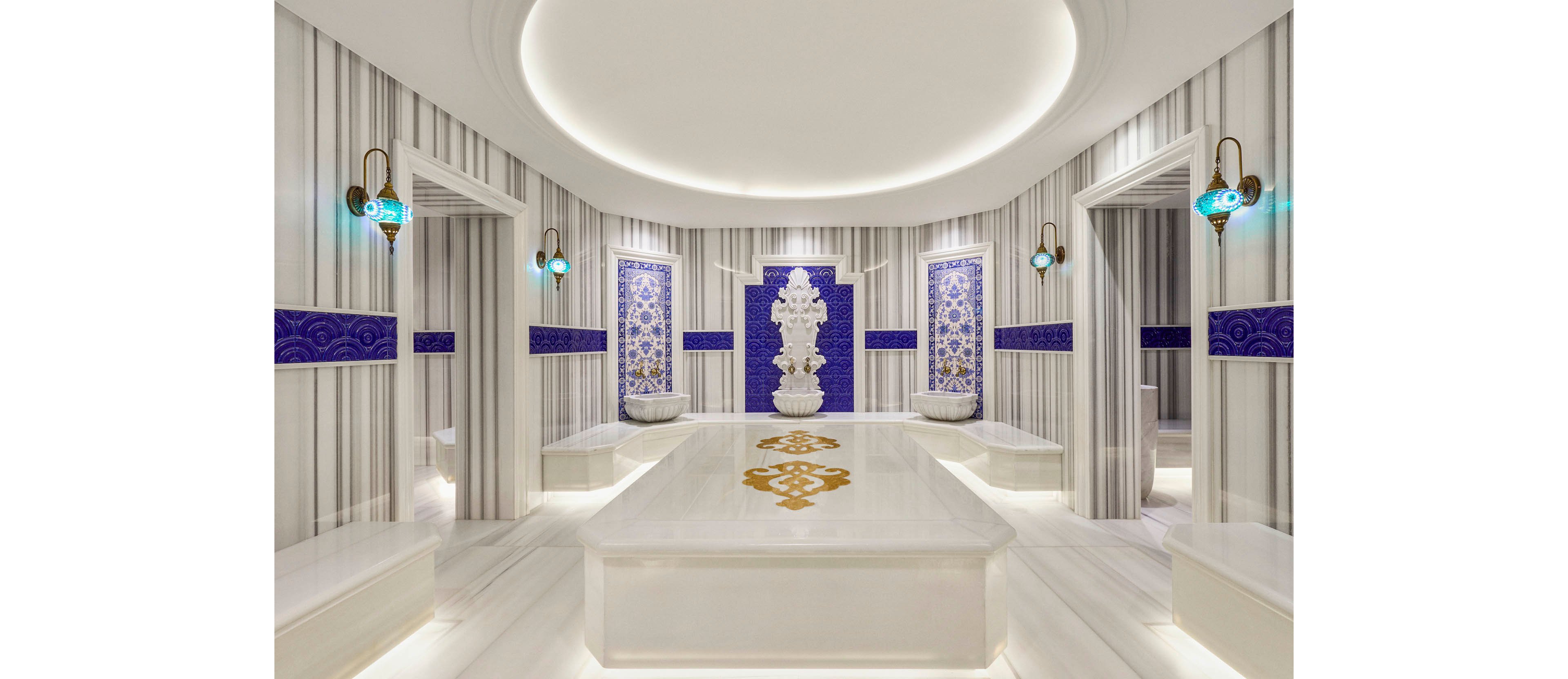 Bright white marble hammam interior with a large central slab dominates the scene. Ornate blue and white tile panels and a decorative fountain create a traditional Turkish bath atmosphere. Wall-mounted brass sconces with turquoise glass add warm accent lighting. The space appears clean, symmetrical, and serene, designed for relaxation and spa rituals.