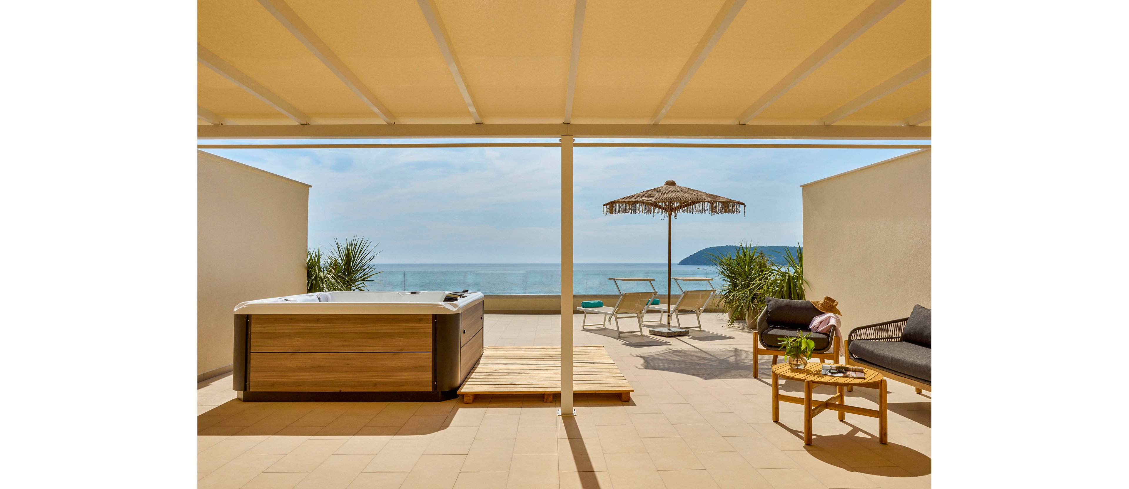 A modern rooftop terrace features a wooden hot tub overlooking a calm ocean horizon. Sun loungers and a straw parasol are positioned toward the water, creating a relaxing vacation setting. On the right, a cozy seating area with a sofa and round table holds drinks and decor items. Warm sunlight, neutral tones, and minimal design emphasize a tranquil, Mediterranean-style atmosphere.