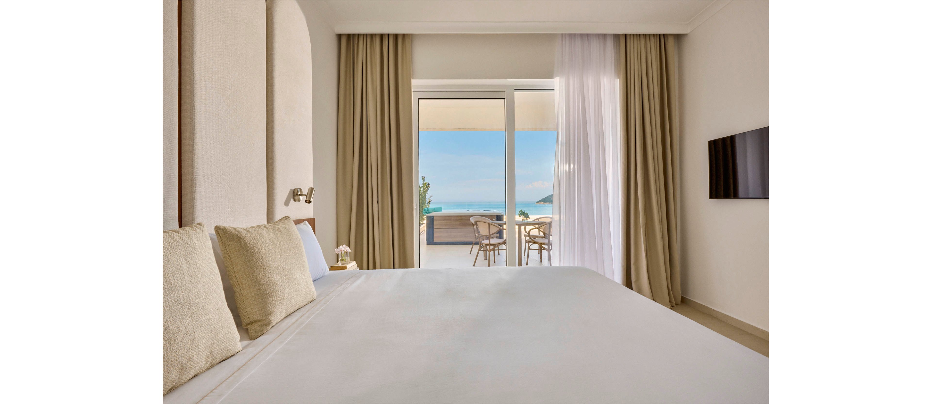 A neatly made bed with light-colored linens and pillows dominates a modern hotel room. Floor-to-ceiling glass doors open onto a balcony with a small table and chairs overlooking the ocean and distant coastline. Neutral walls and beige curtains create a calm, minimalist atmosphere, complemented by a wall-mounted television. Soft natural light fills the space, emphasizing the airy and relaxing seaside setting.