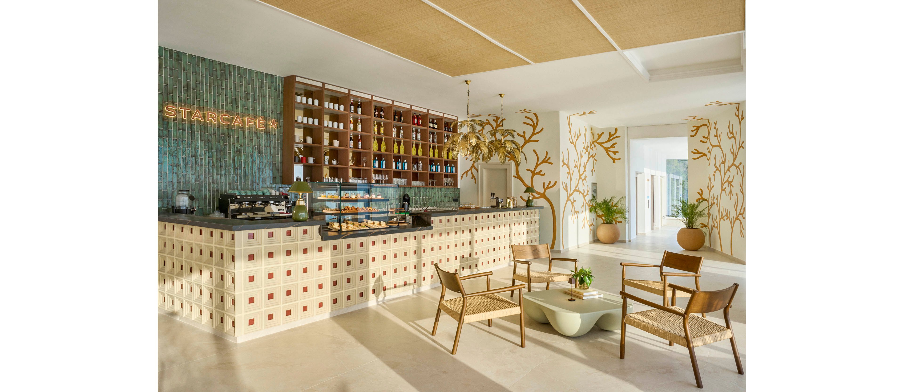 Stylish café interior features a long tiled counter with the illuminated word STARCAFÉ on the back wall. Shelving behind the bar displays bottles and products, while a glass case showcases pastries. In the foreground, woven chairs surround a low sculptural coffee table, and decorative plants and wall art add a natural touch. Sunlit corridors and neutral tones create an airy, welcoming atmosphere.