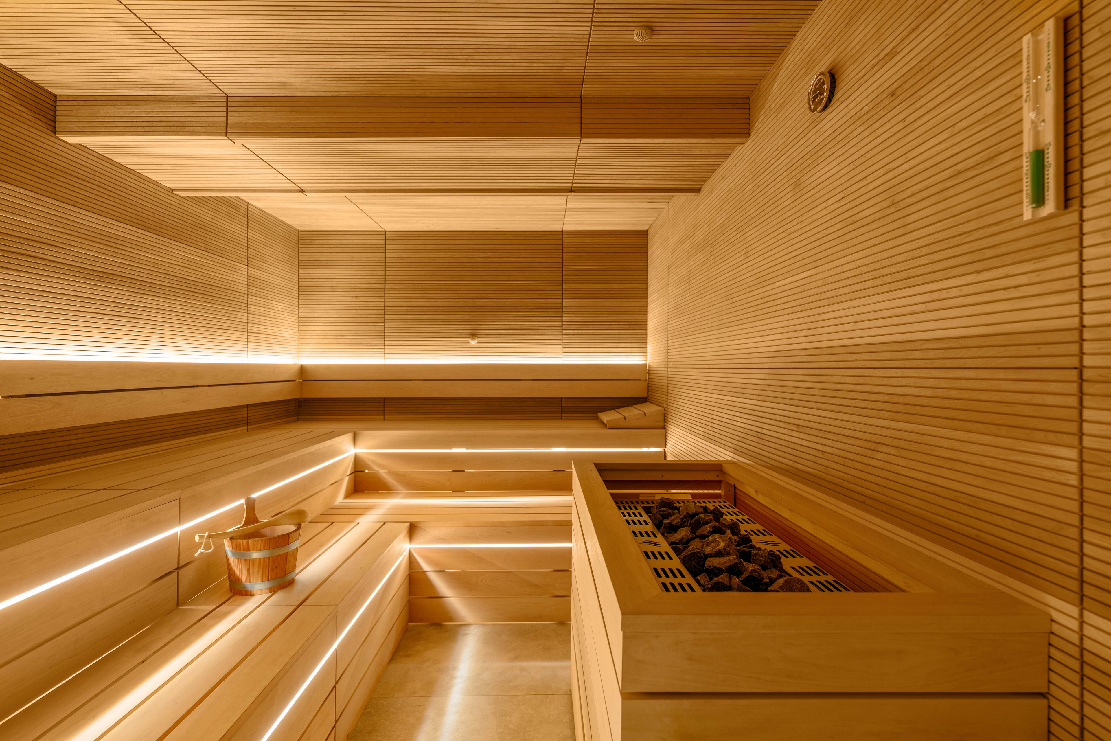 Contemporary sauna room featuring smooth wooden benches and walls is illuminated by integrated warm strip lighting. A heater filled with sauna stones is positioned in the foreground, emphasizing the spa-like environment. A wooden bucket and ladle sit on the bench, suggesting relaxation and traditional sauna rituals. The overall design is minimalistic, clean, and inviting, with a golden, cozy glow throughout the space.