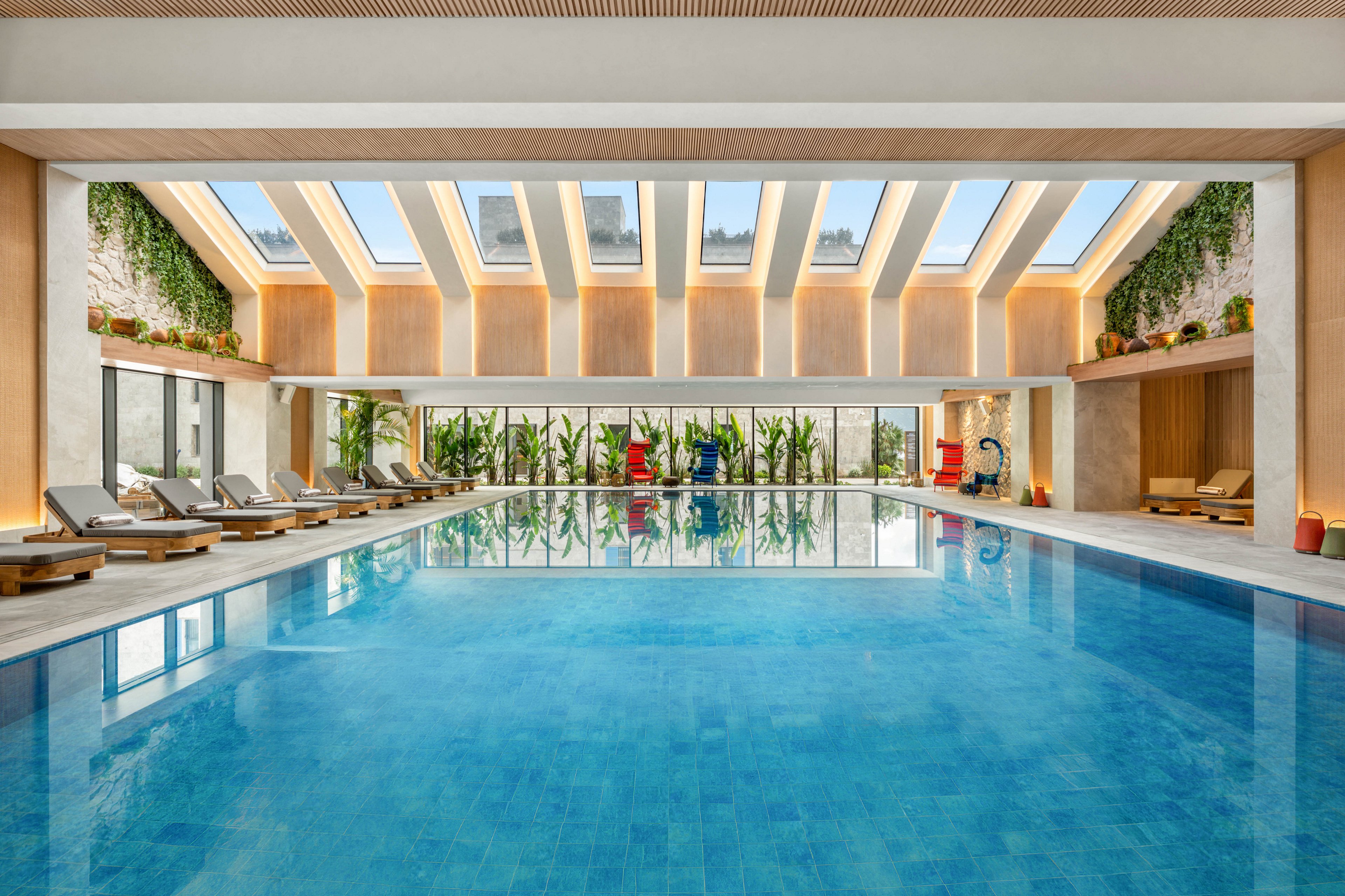 A large indoor swimming pool occupies the center of a modern, light-filled room. Sunlight streams through multiple skylights in the ceiling, reflecting on the clear blue water. Lounge chairs line both sides of the pool, accompanied by potted plants and greenery along the walls. The overall design feels contemporary and relaxing, with warm tones and natural elements creating a spa-like atmosphere.