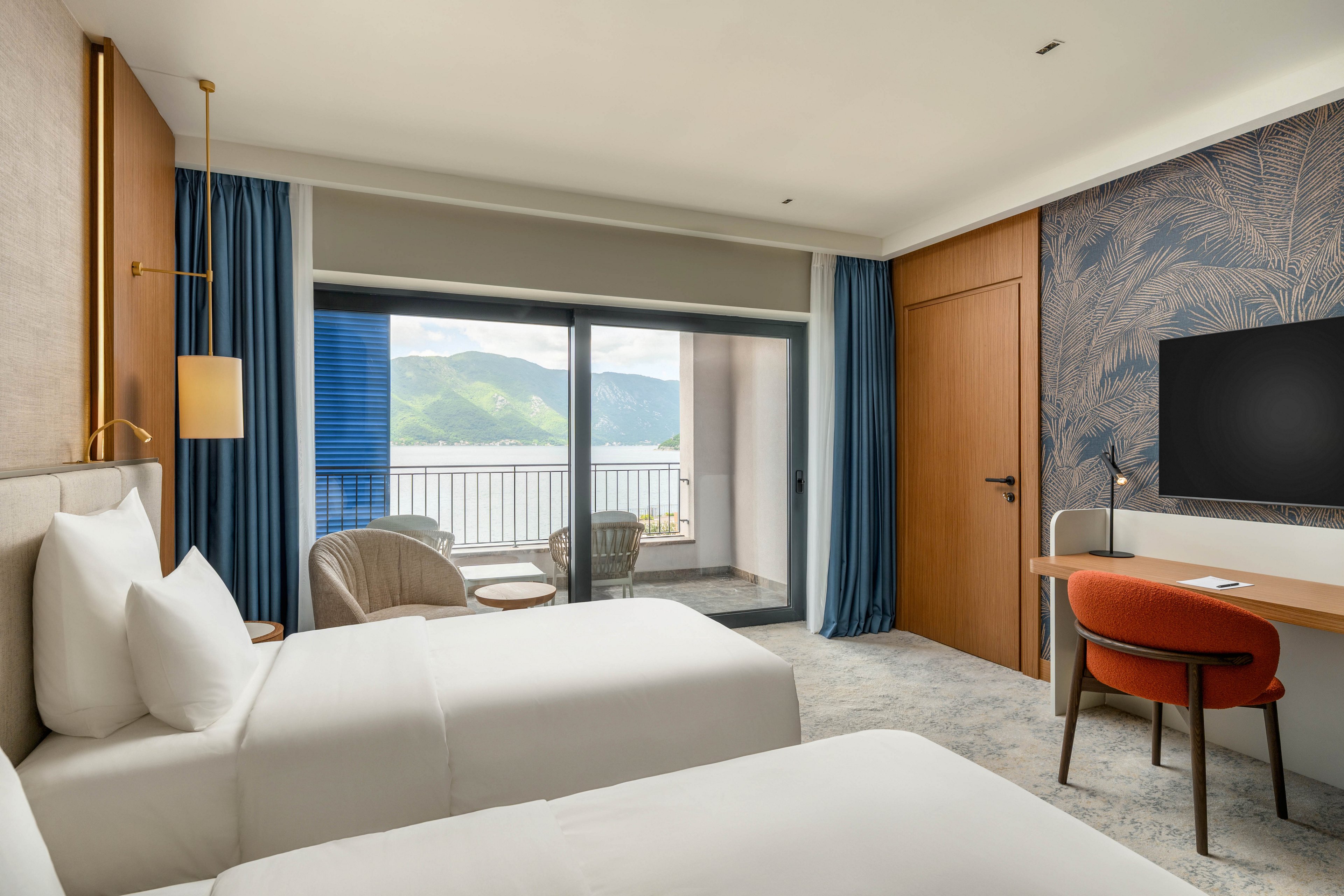 Contemporary hotel room featuring two neatly made twin beds faces a balcony with scenic mountain and water views. A seating area with woven chairs and a small table is positioned near the sliding glass doors. On the right, a desk with an orange chair and a large flat-screen TV sits against a patterned accent wall. Soft neutral tones with blue curtains and warm wood accents create a calm, inviting atmosphere.